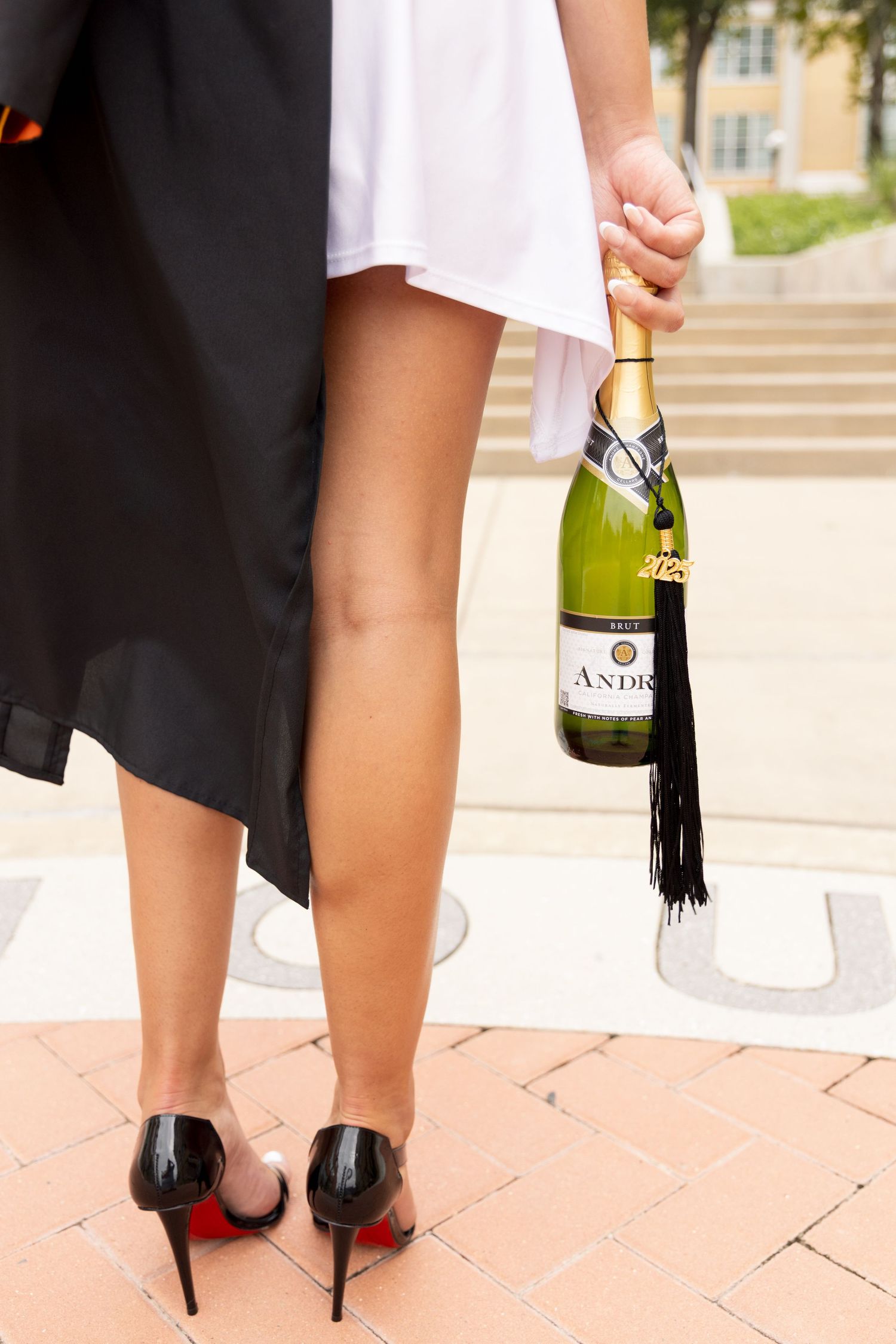 Graduate in a white dress and black high heels holds a champagne bottle with a black tassel tied to its neck on a brick campus walkway.