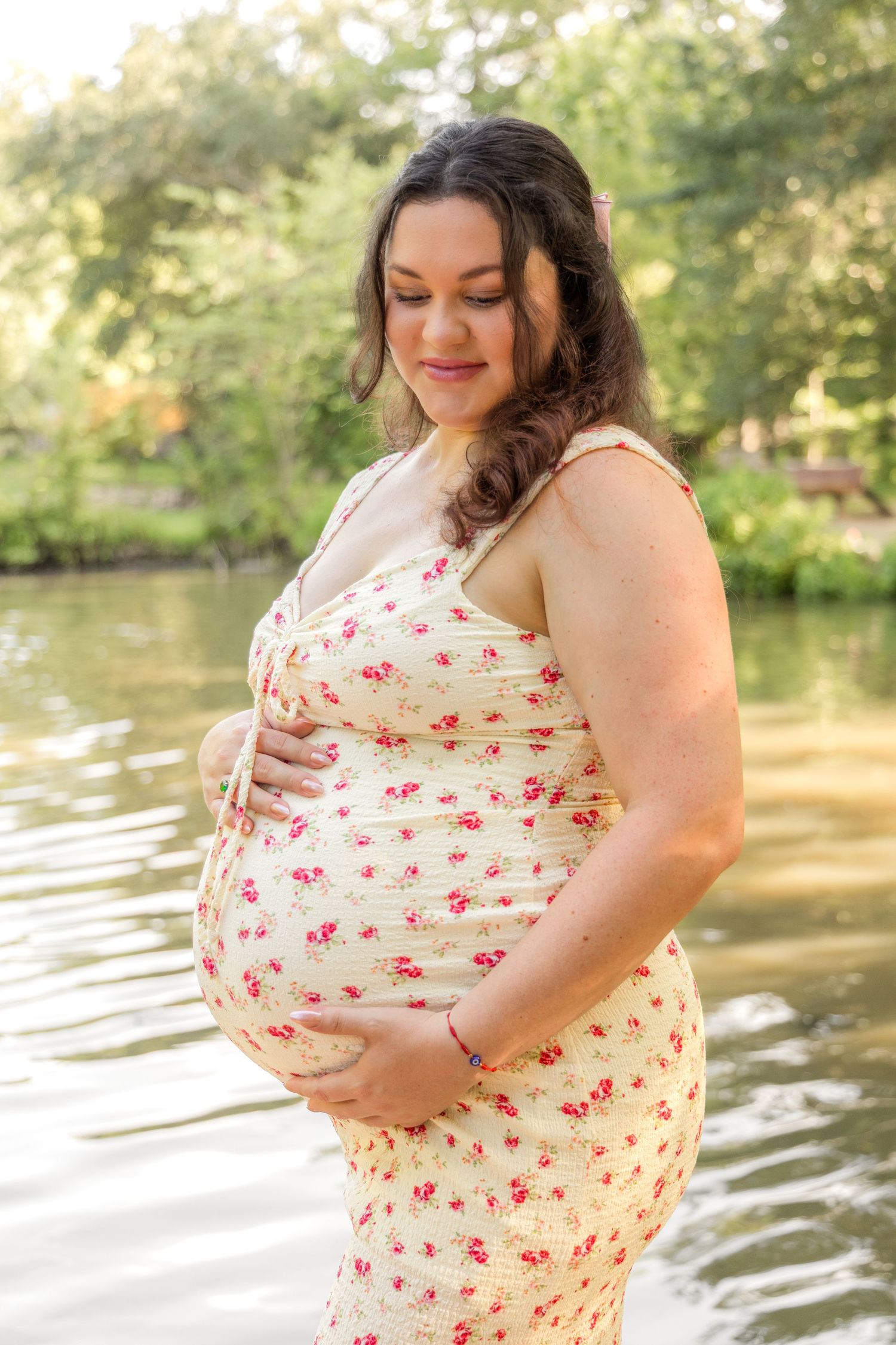 Pregnant woman in a cream dress with red floral print cradling her belly beside a calm lake surrounded by greenery.