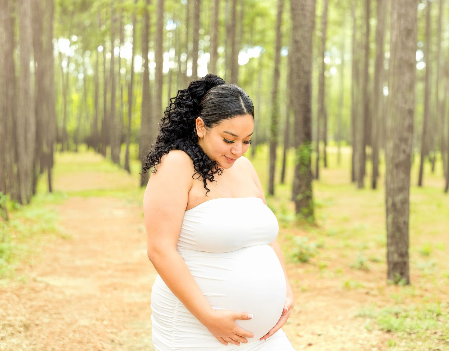 Expecting mother in a white dress gently cradles her baby bump on a sunlit forest path lined with tall pines.