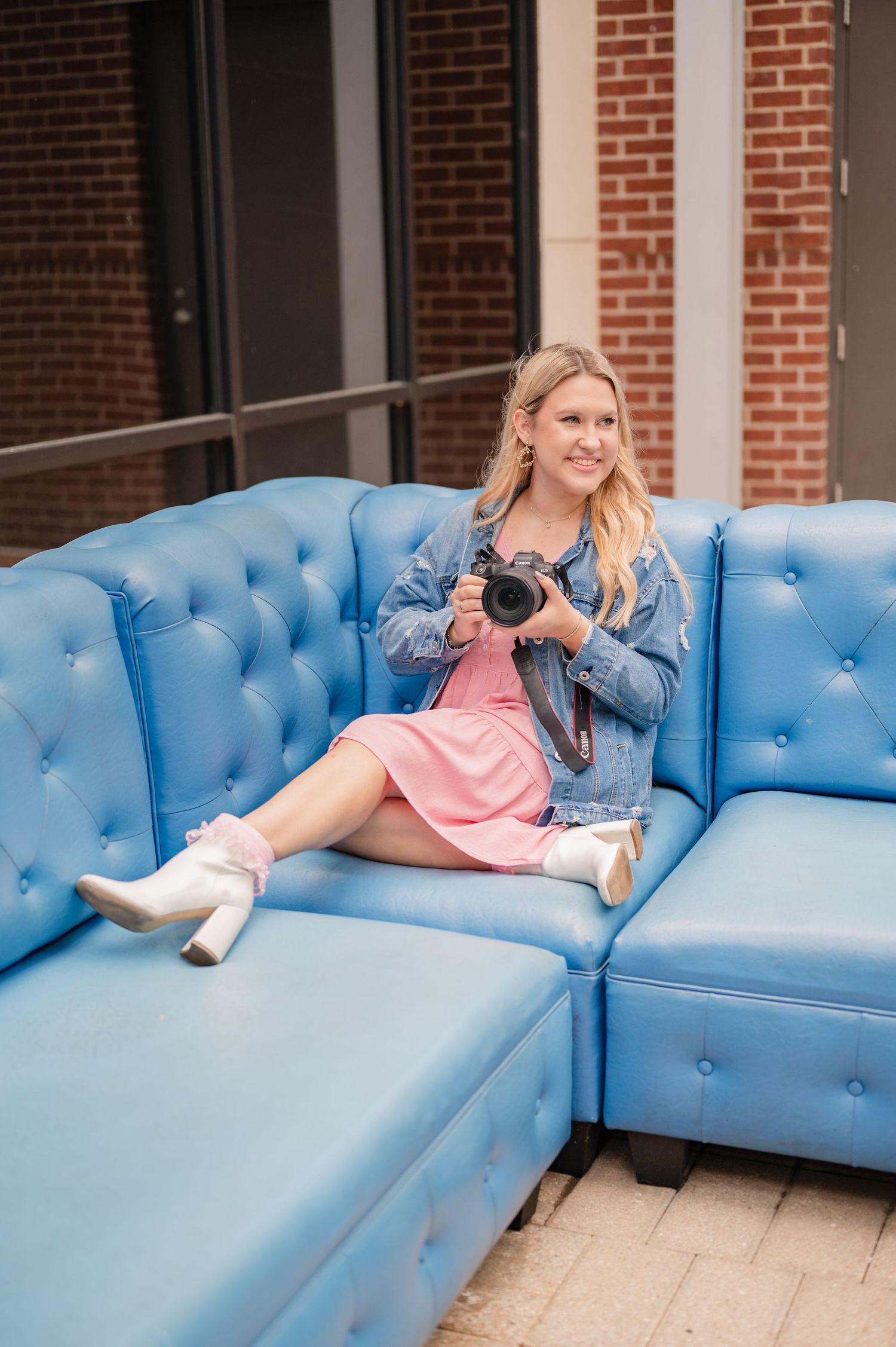 Blonde woman in a denim jacket and pink dress sits on a blue tufted sofa holding a camera outside a brick building.