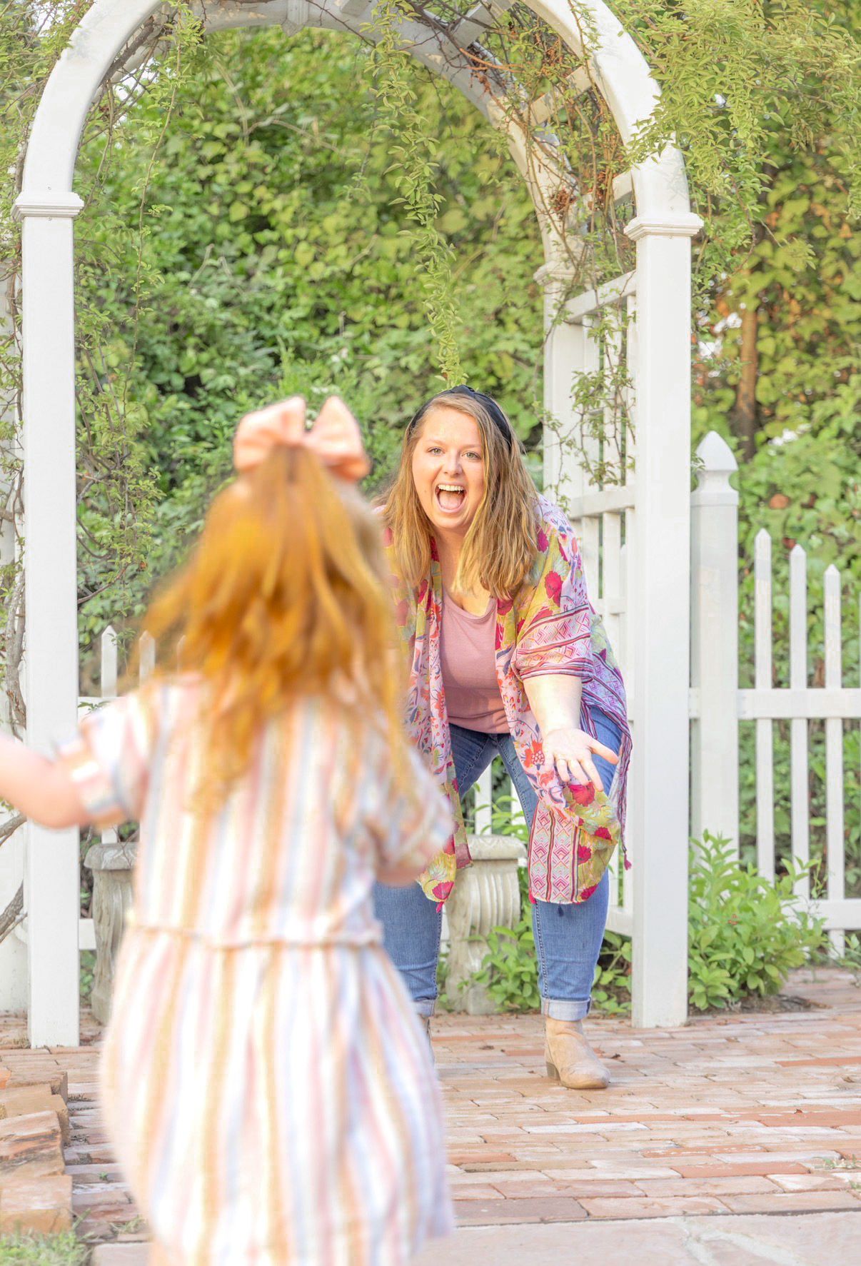 Laughing woman in a colorful scarf crouches with arms open beside a white garden arbor, greeting a little girl running toward her.
