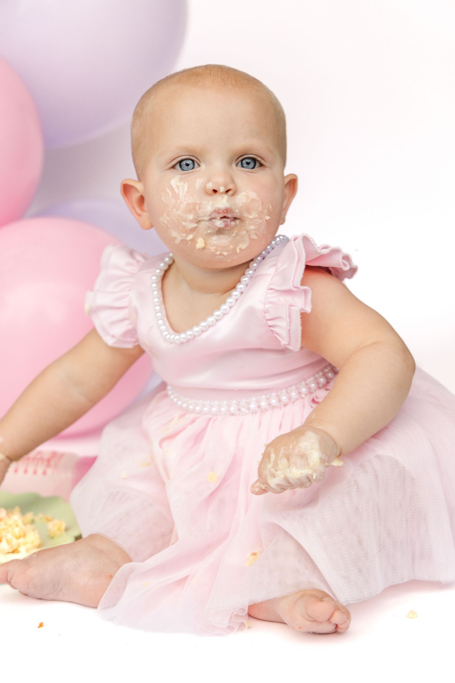 Baby girl in a pink tulle dress and pearl necklace with cake smeared on her face and hands, seated near pastel balloons.