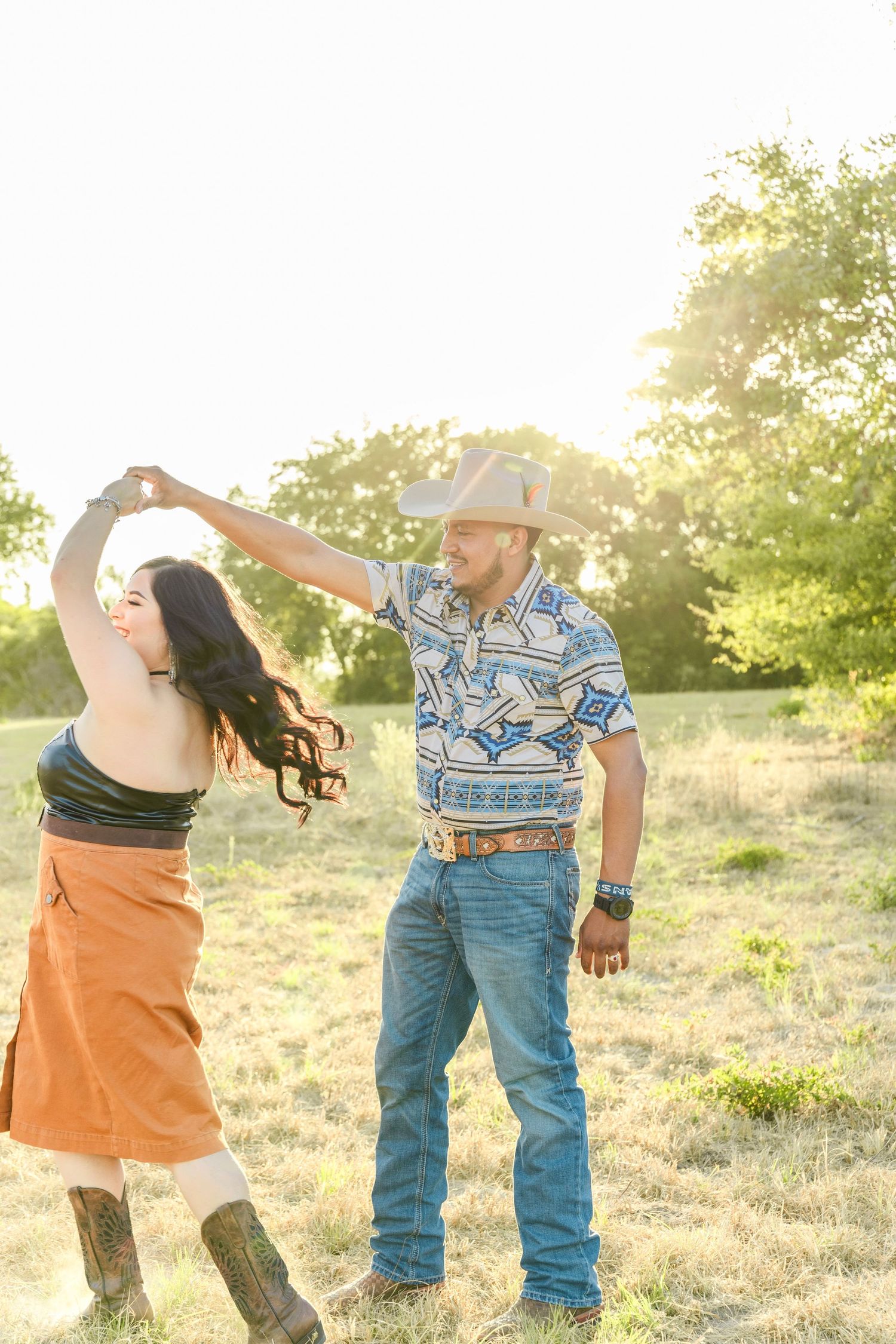 Couple dances in a sunny grassy field as he twirls her, he in a cowboy hat and western shirt, she in boots.