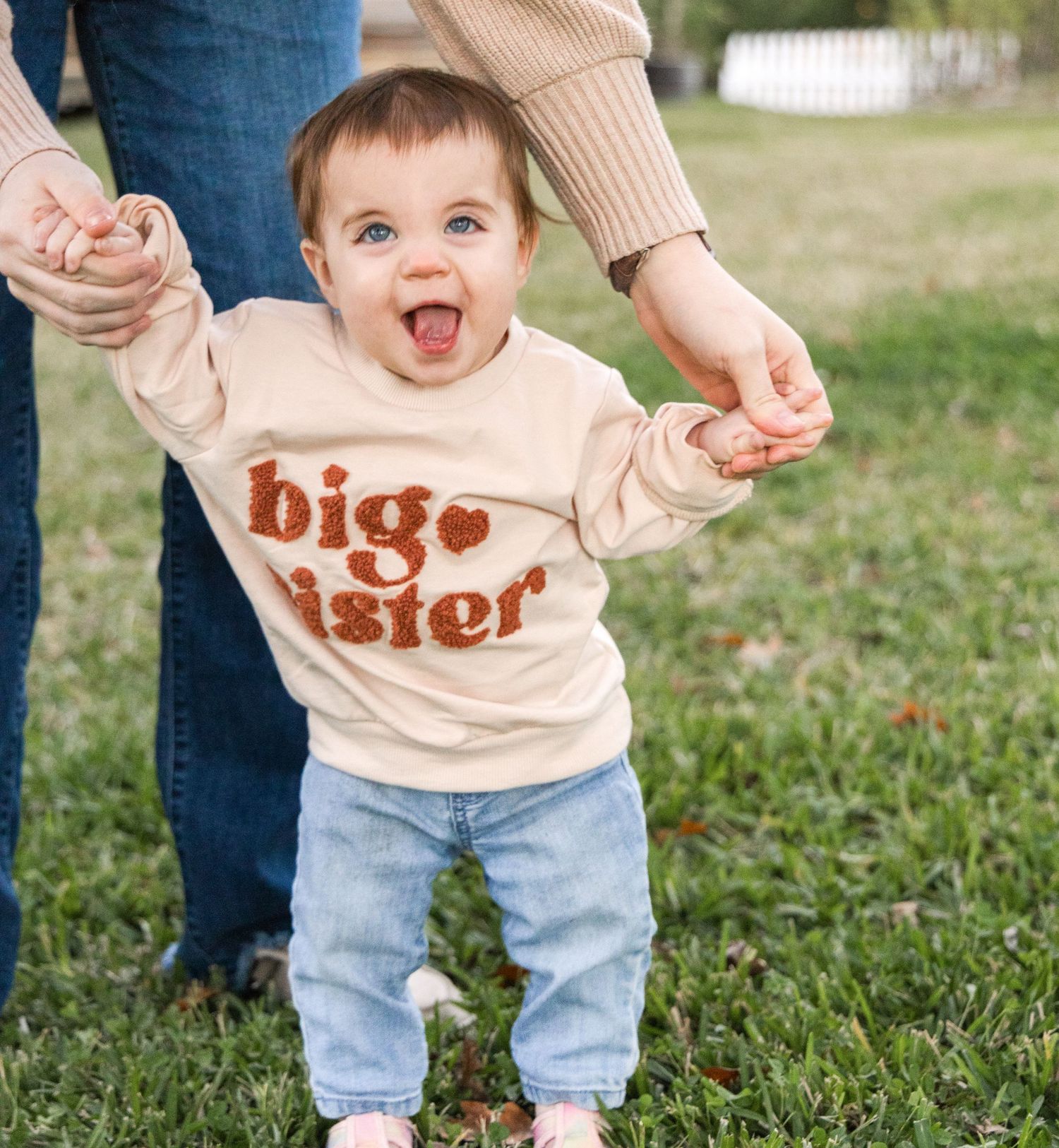A laughing toddler in a big sister sweatshirt and jeans holds hands with a parent while standing on green grass.