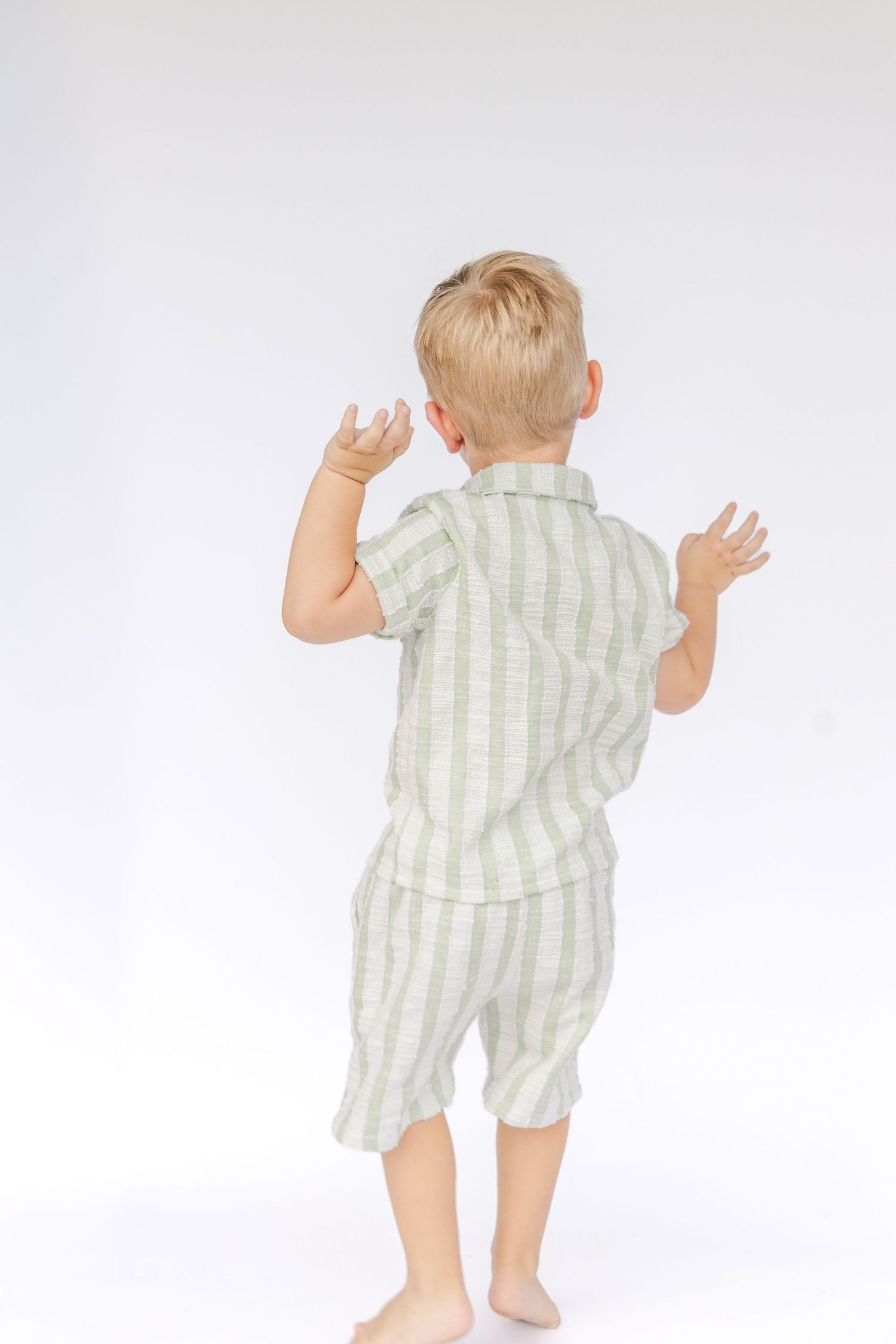 A barefoot toddler with blond hair wearing a striped green and white shirt and shorts raises both hands, photographed from behind on white.