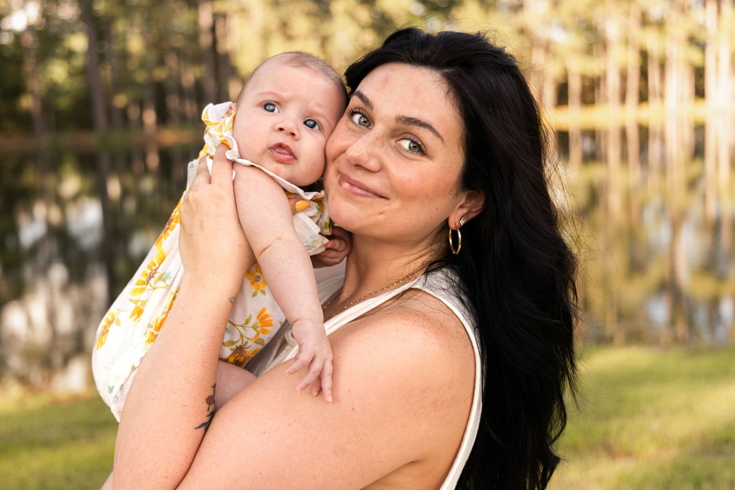 Dark-haired mother smiling gently while cradling her baby in a yellow floral outfit in front of a lake and trees.