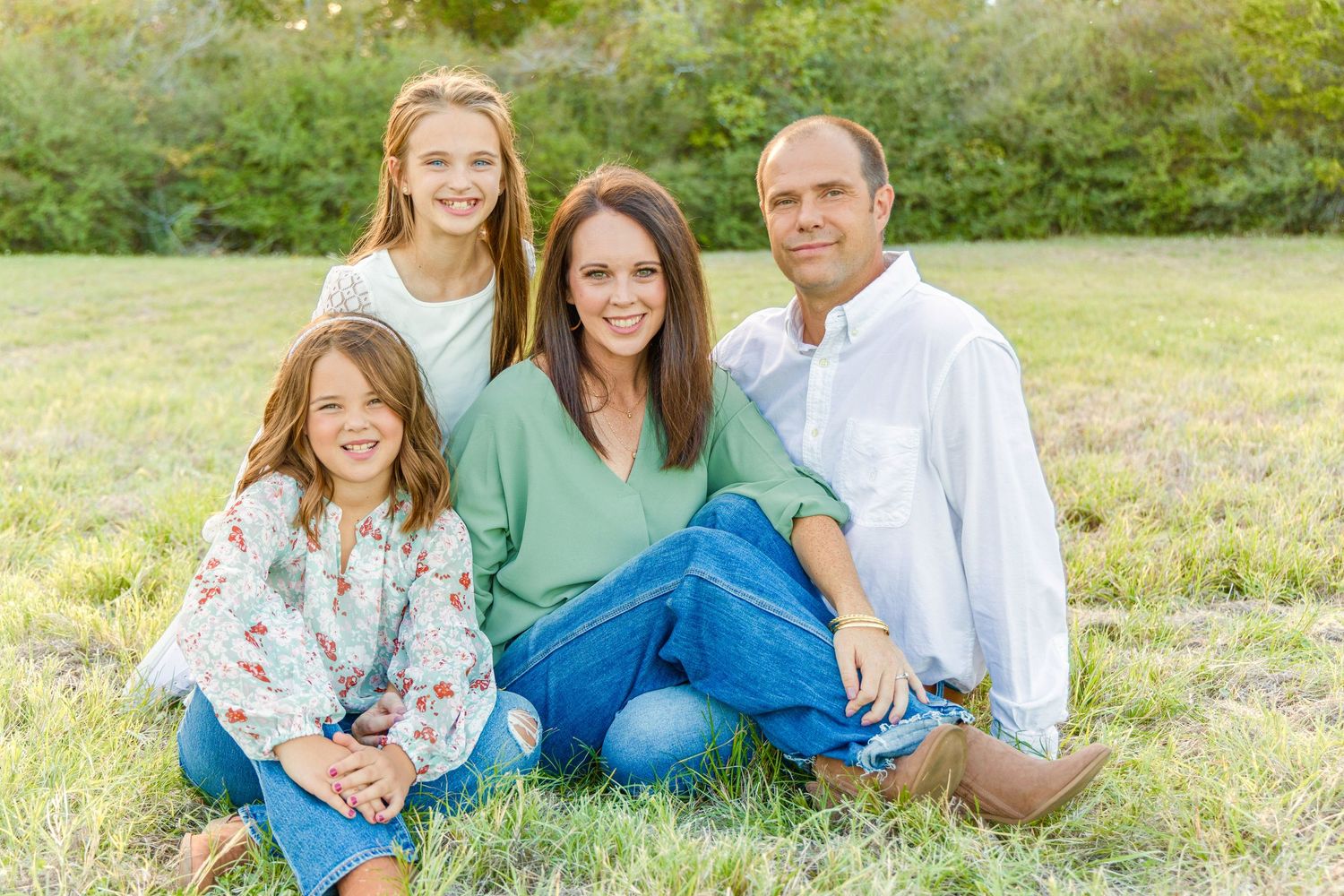Family of four sits together on a grassy field, with smiling parents and two daughters posing for a warm outdoor portrait.