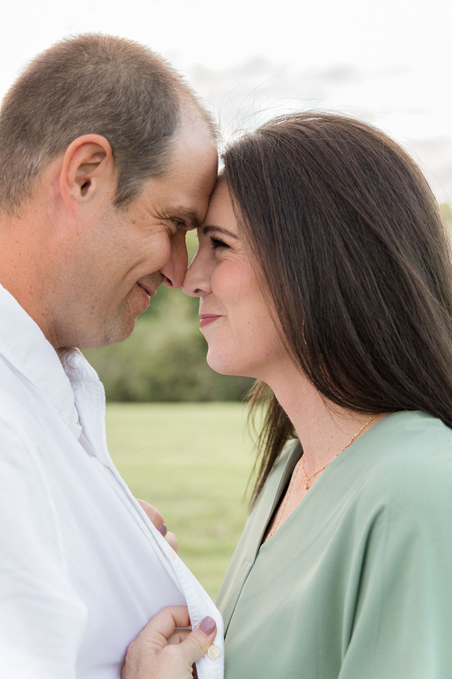 Smiling couple touching foreheads in an affectionate embrace outdoors on a grassy field during an engagement photo session.
