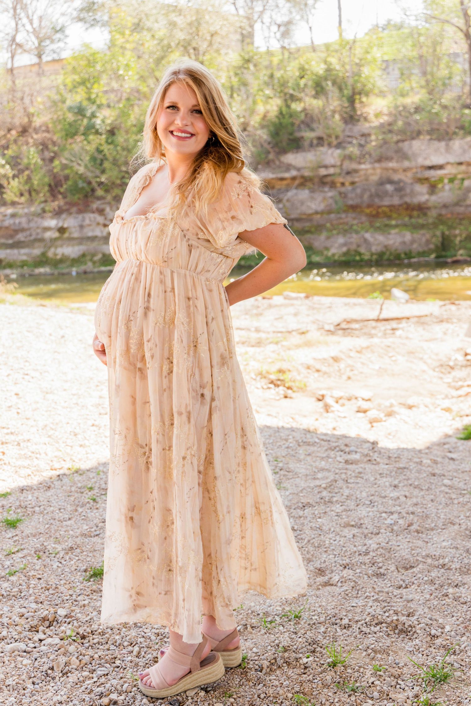 Smiling pregnant woman in a flowing cream floral dress and wedge sandals standing on a gravel creek bed in sunlight.