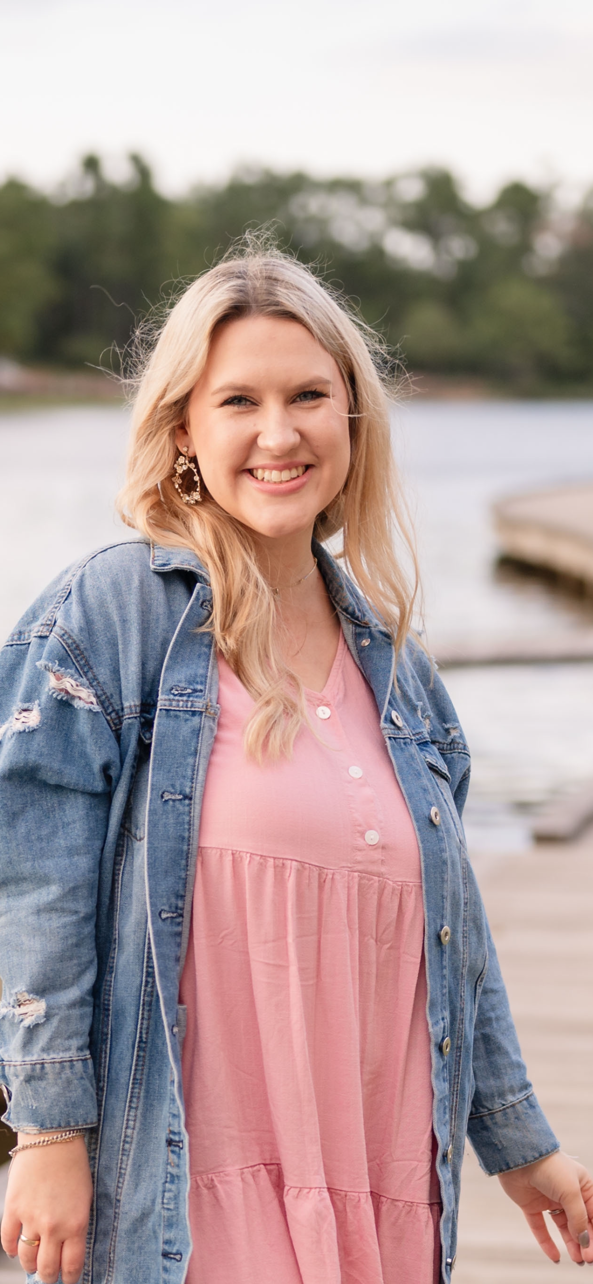 Smiling woman with long blonde hair in a pink dress and denim jacket stands near a lake and wooden dock.