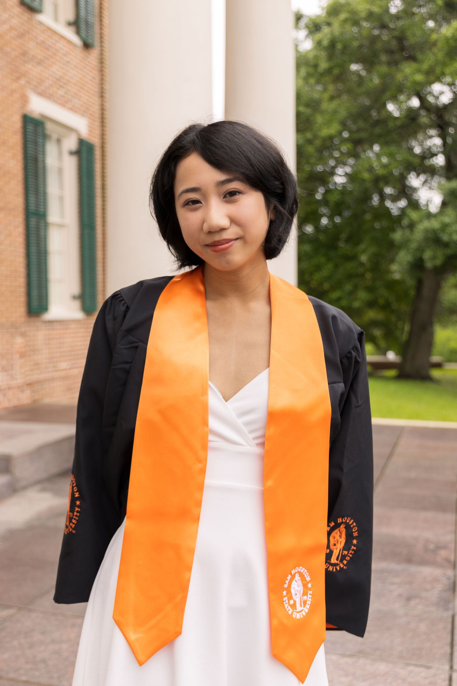 A young graduate with short dark hair wears a white dress, black gown, and orange stole in front of white columns and a brick building.