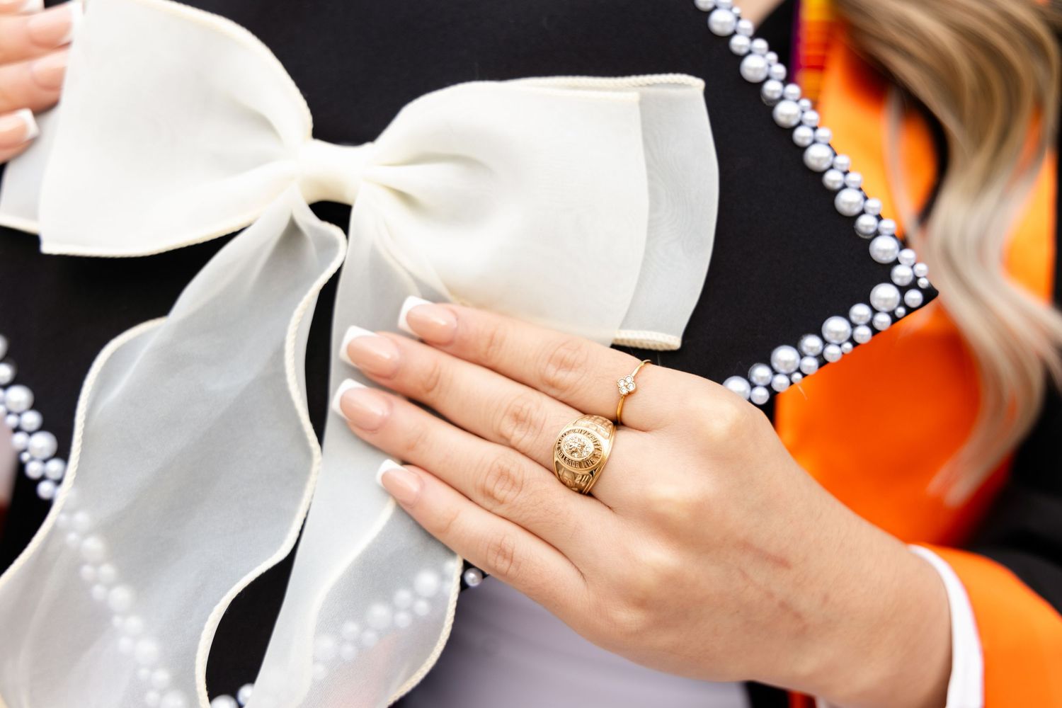 Close-up of a woman's manicured hand with rings resting on a large white bow and pearl-trimmed black jacket with orange accents.