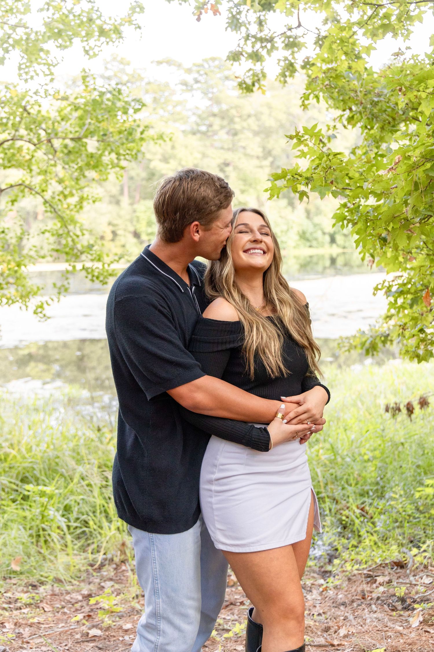 Smiling couple embracing outdoors as he kisses her cheek, with a calm lake and green trees behind them.