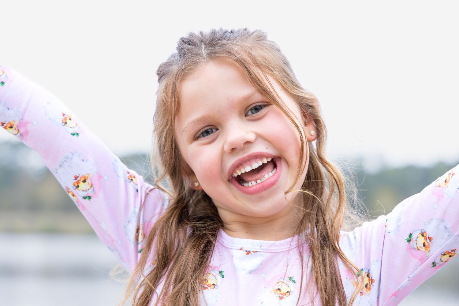 A joyful little girl in a floral pink shirt laughs with her arms stretched wide outdoors near a calm lake.