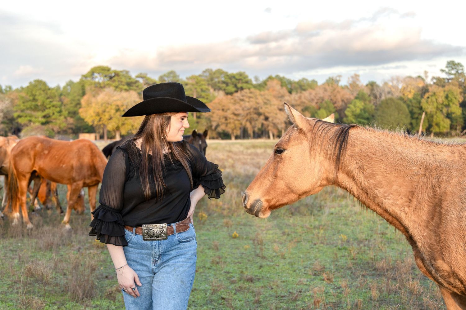 Woman in a black cowboy hat and sheer black blouse stands in a grassy pasture beside a palomino horse at golden hour.