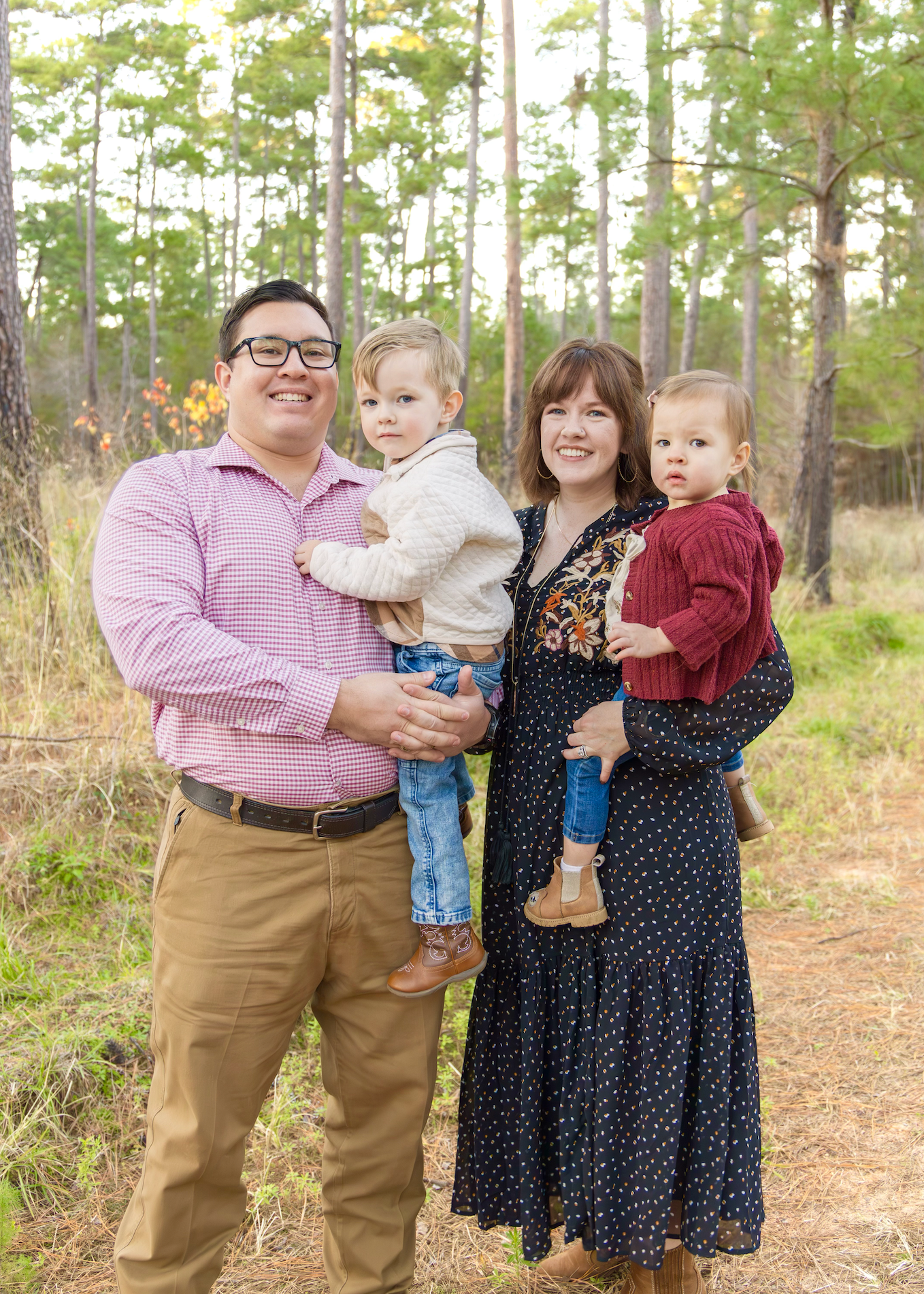 Smiling family of four poses in a pine forest, with parents holding two young sons during an outdoor portrait session.