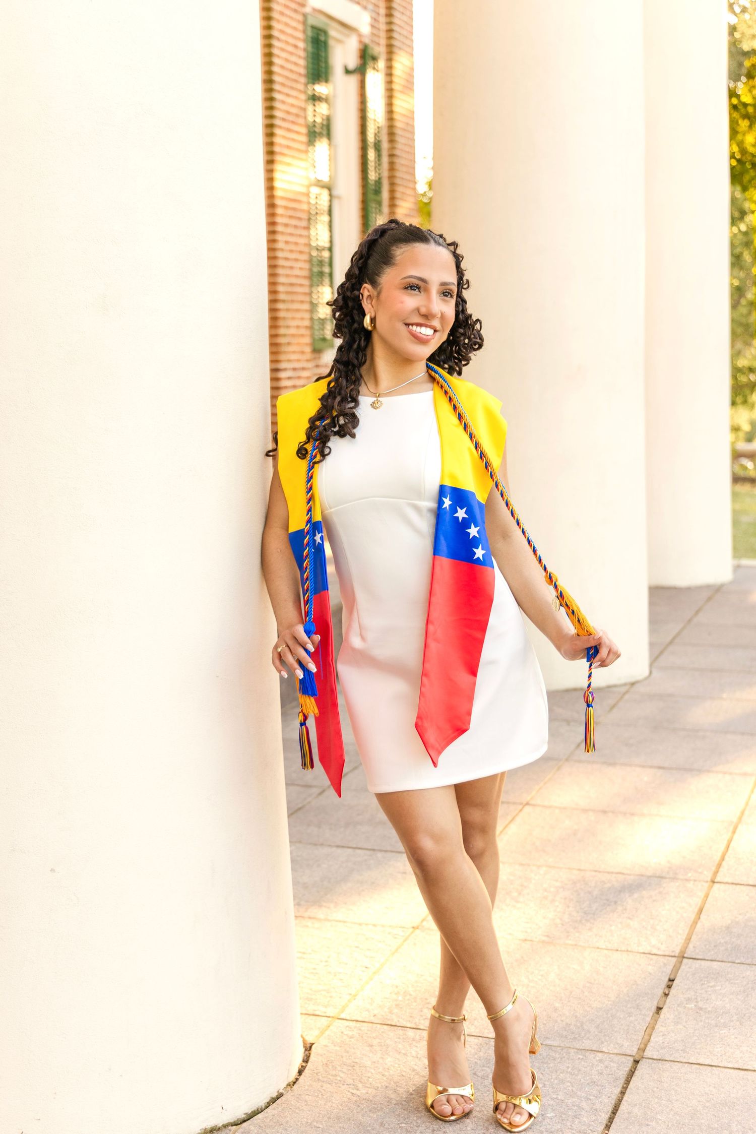Smiling graduate in a white dress wears a Venezuelan flag stole and honor cords while posing beside white columns on campus.