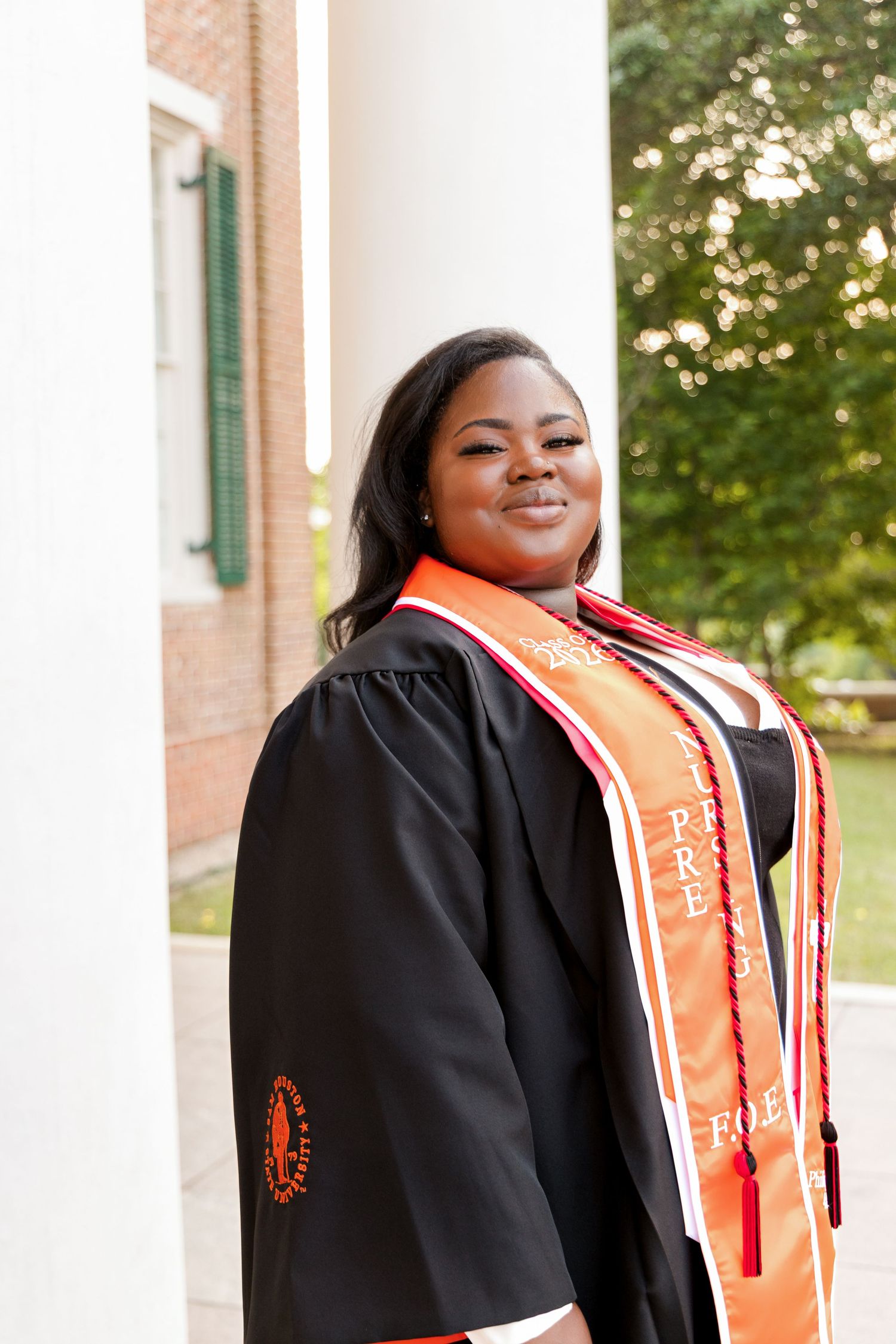 Smiling graduate in a black gown and orange honor stole poses between white columns beside a brick building on campus.