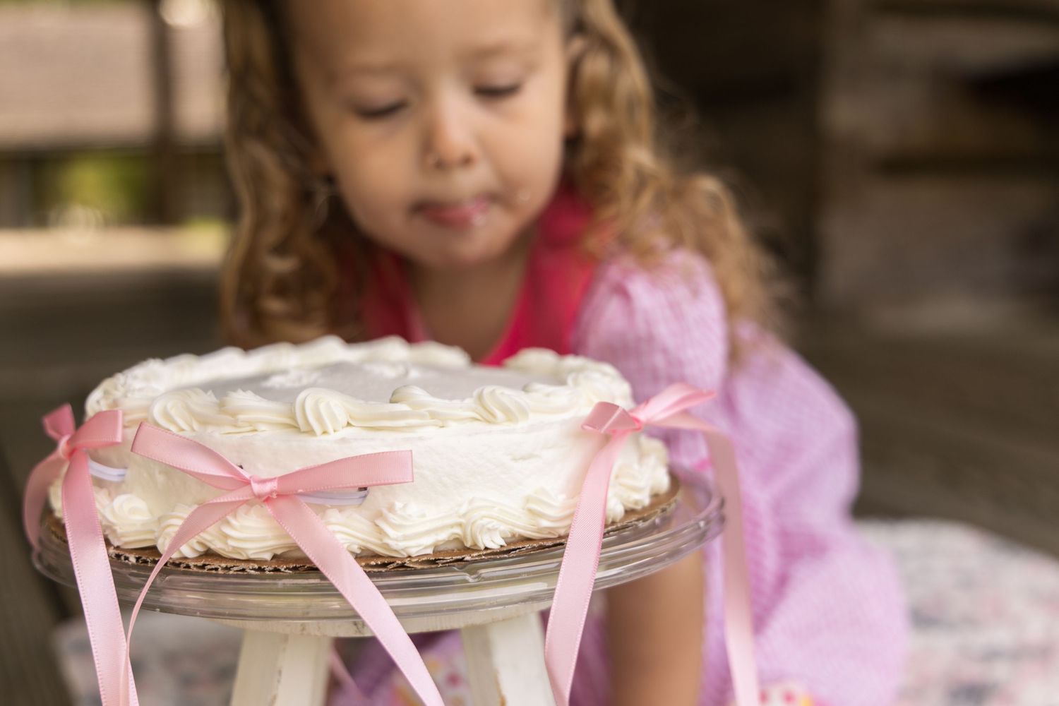 A curly-haired toddler in a pink dress gazes at a white frosted cake with pink ribbons on a glass cake stand.