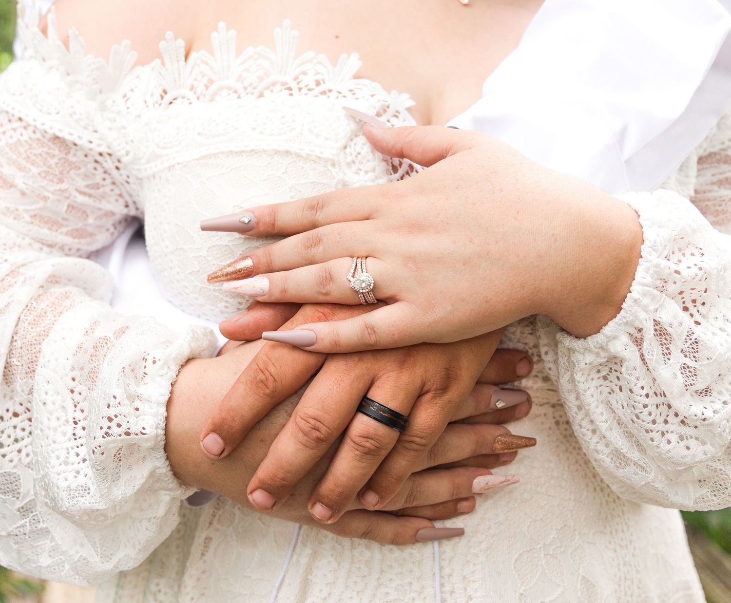 Close-up of a bride and groom's clasped hands showing a diamond engagement ring and a black wedding band with a lace dress.