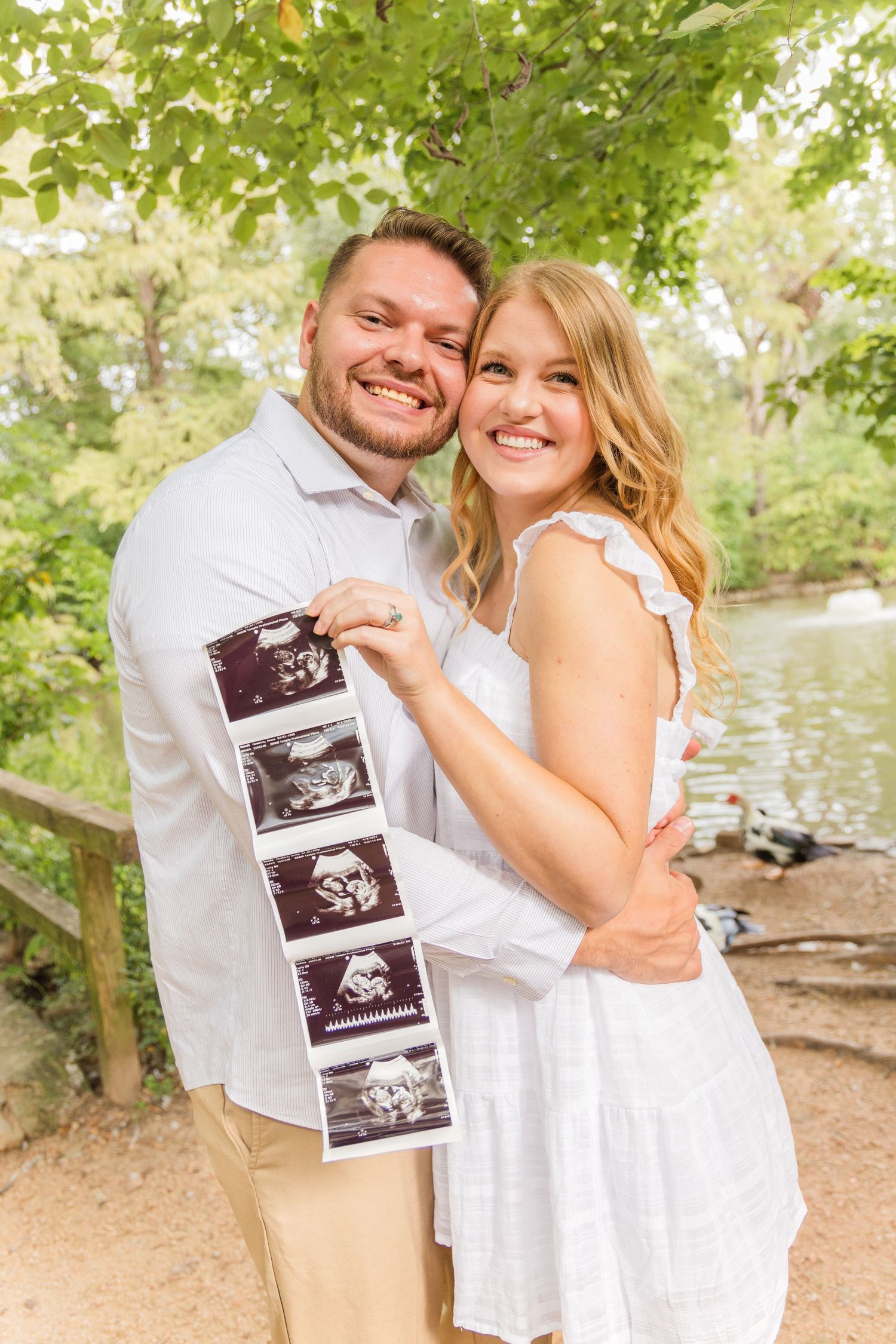 Happy expecting couple in white outfits hug beside a lake while holding a strip of ultrasound photos in a sunny park.