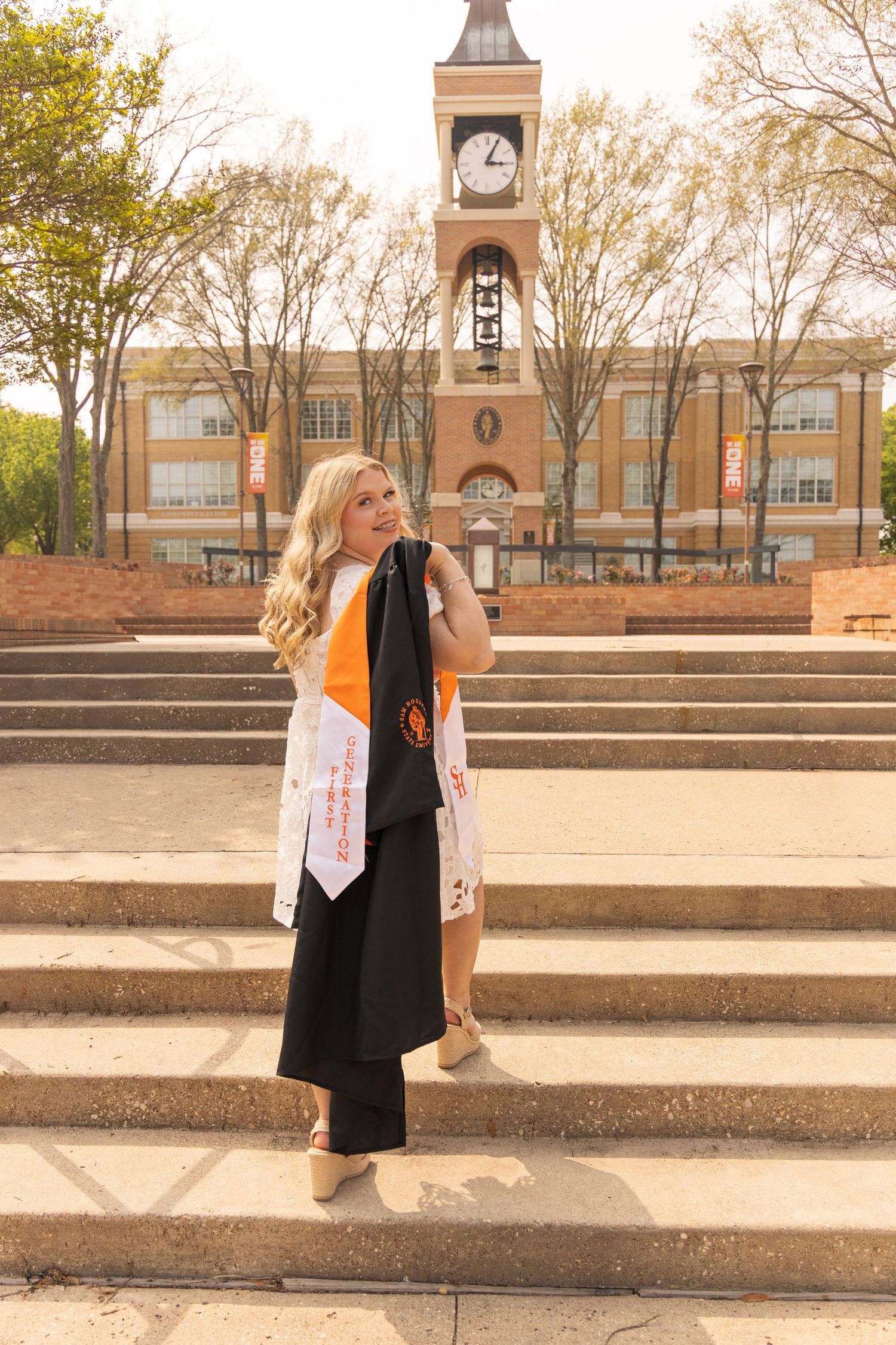 A smiling graduate in a black gown and orange stole poses on brick steps in front of a historic campus clock tower.
