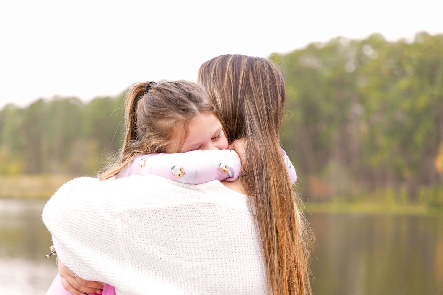 A child in a pink floral top hugs a long-haired girl in a white sweater by a peaceful lake and trees.