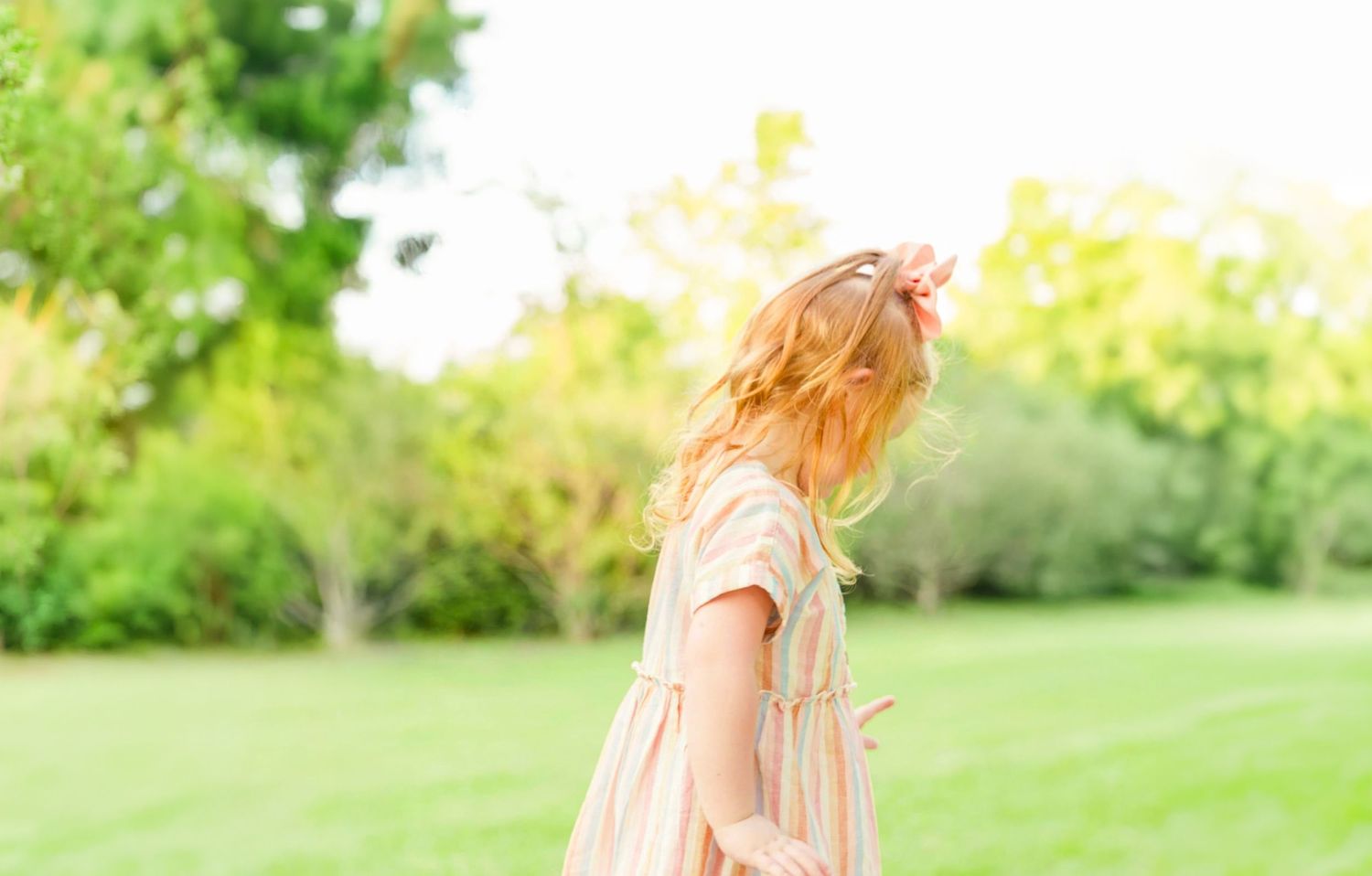 A young girl with wavy blonde hair in a pastel dress twirls in a sunny green park surrounded by soft, blurred trees.