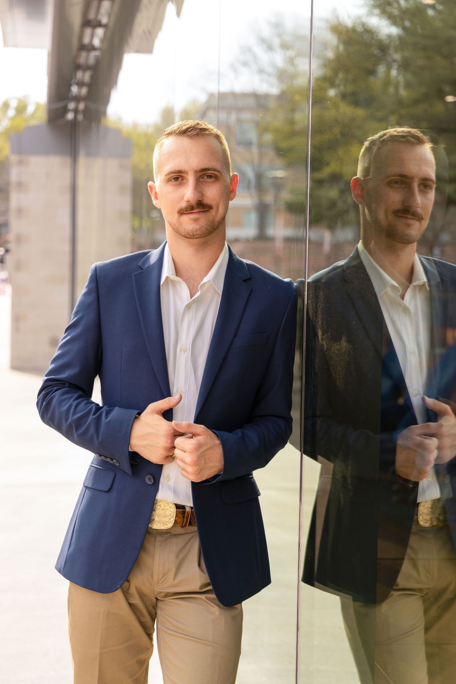 Man in a navy blazer, white shirt and khaki pants poses beside a glass wall with his reflection visible.