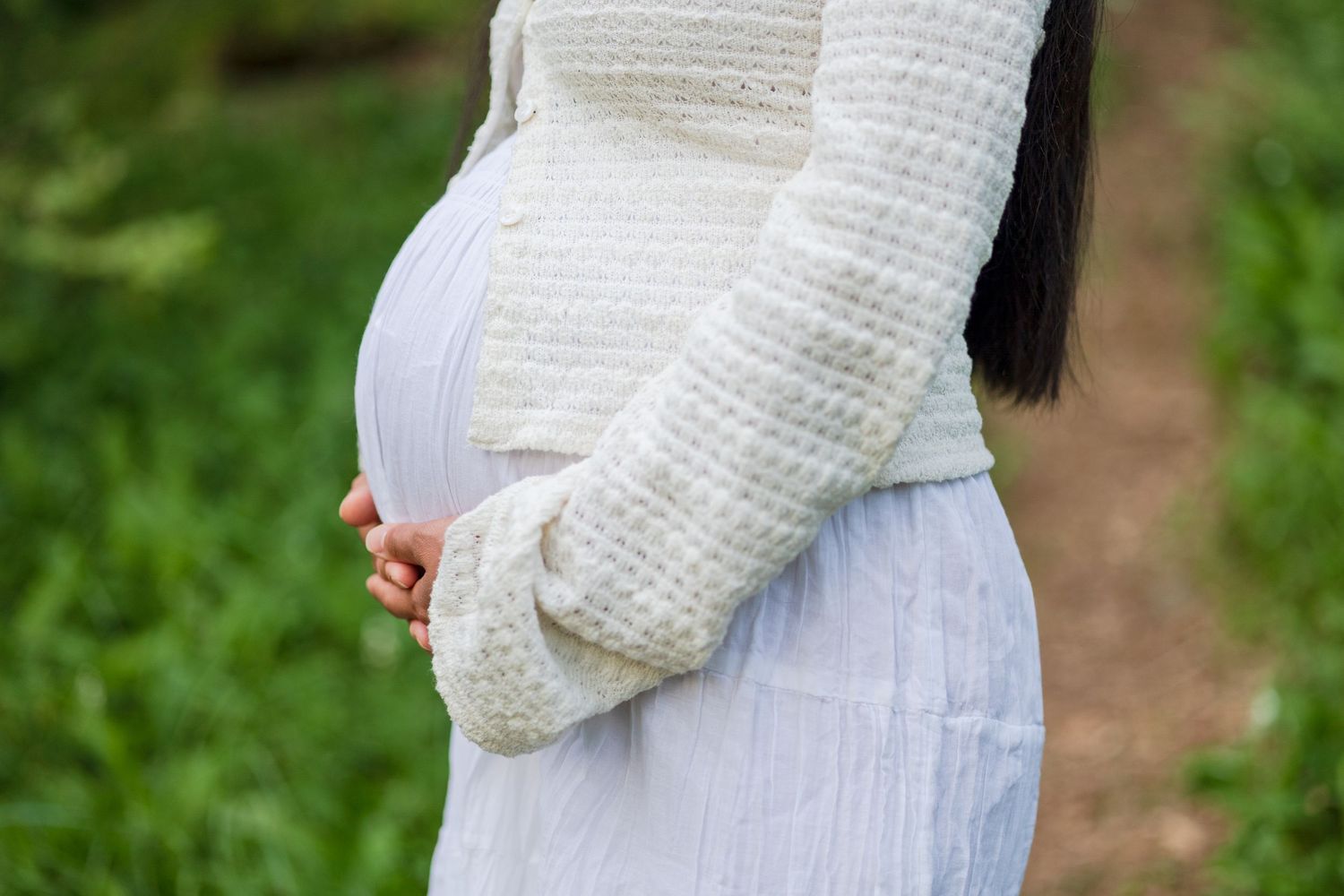 Pregnant woman in a white knit cardigan and flowing white skirt cradling her baby bump outdoors near green foliage.