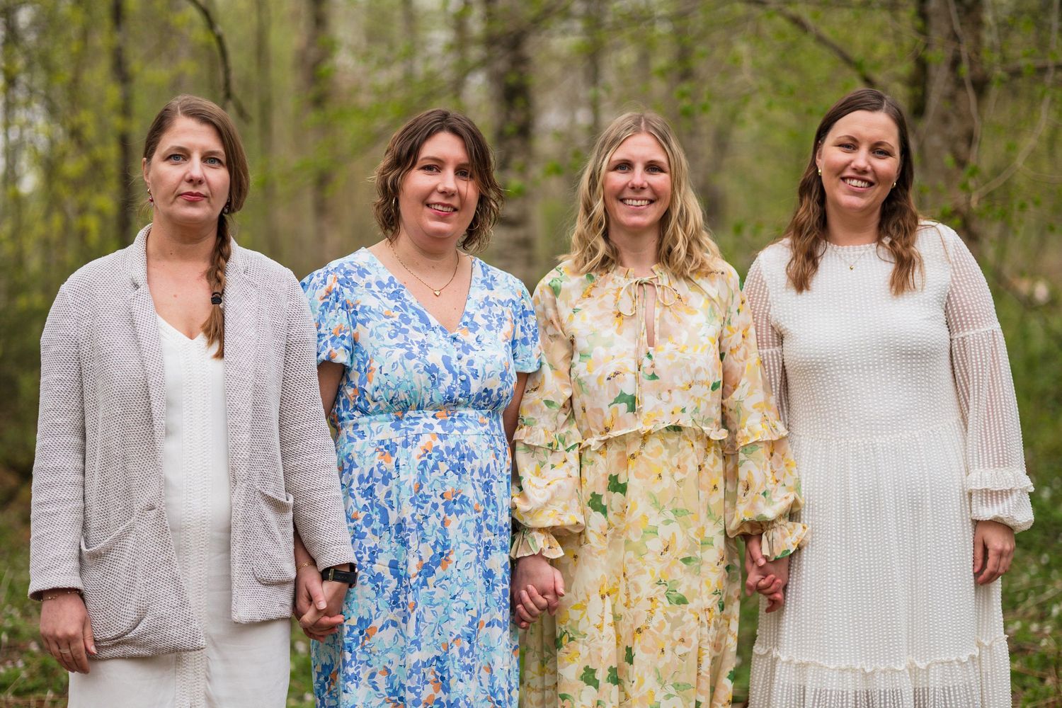 Four smiling women in light spring dresses and cardigans stand together holding hands in a green forest.