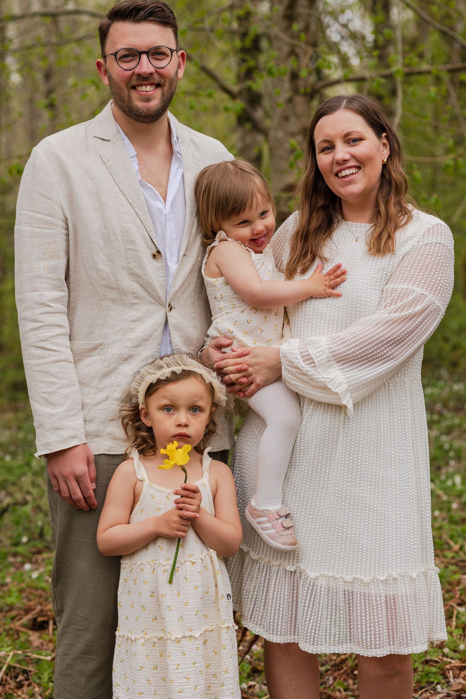 Smiling family of four in light summer outfits poses in a spring forest, with a girl holding a yellow flower.