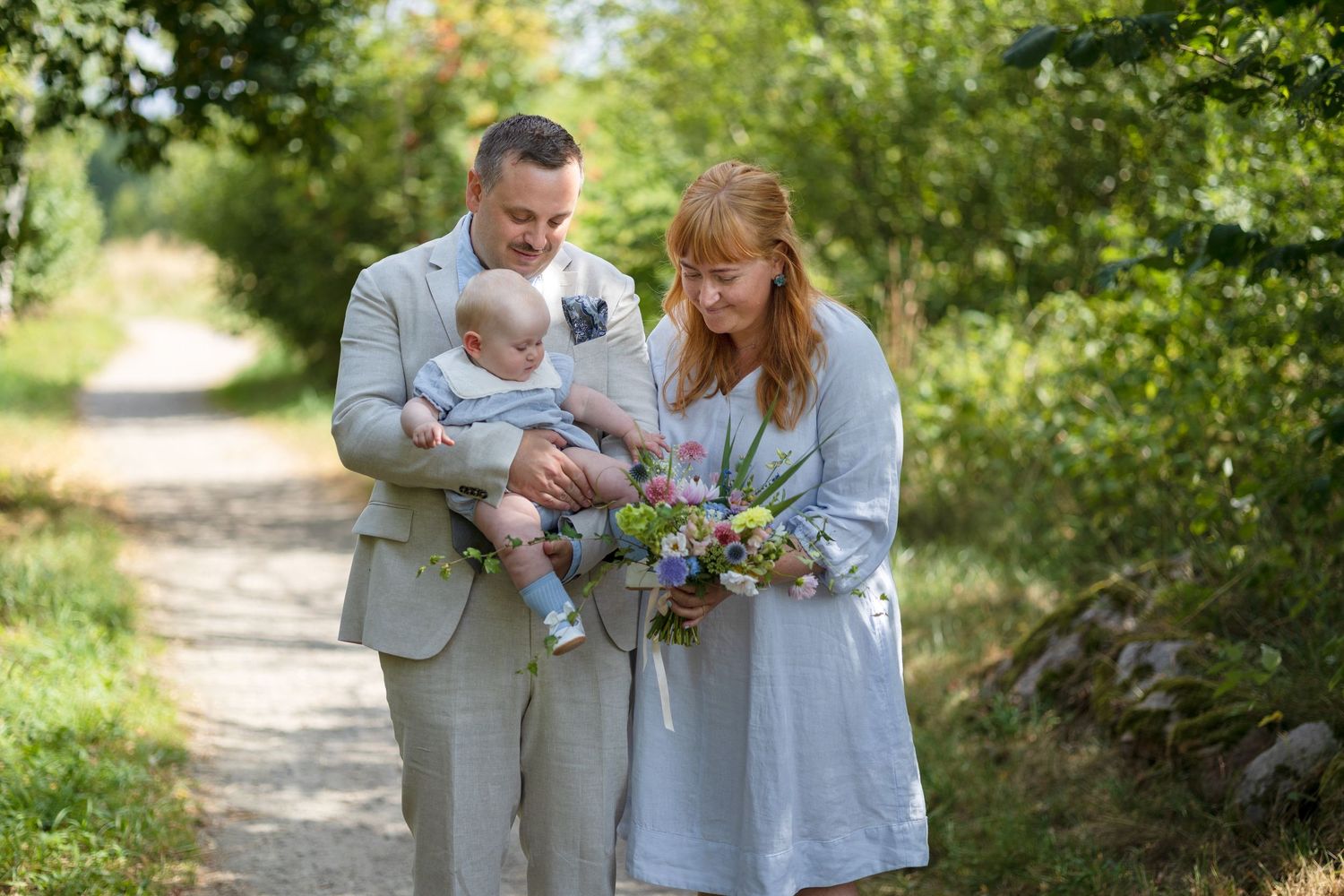 A smiling couple holds their baby and a colorful wildflower bouquet while standing on a sunny path surrounded by greenery.