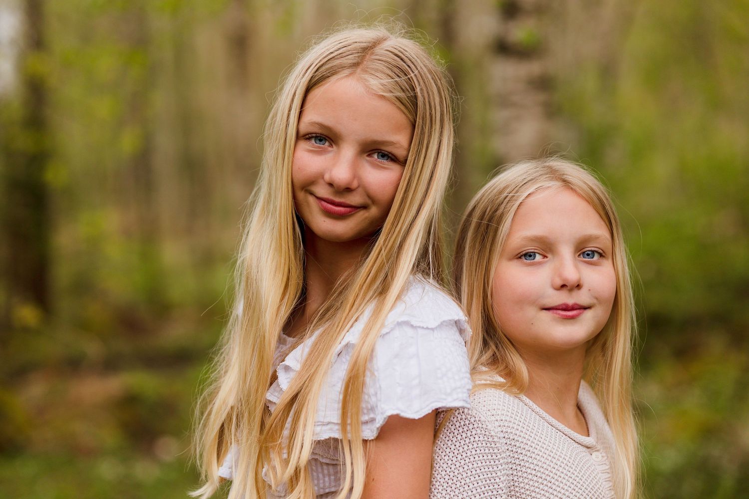 Two smiling blonde sisters pose back to back in a softly blurred green forest in spring.