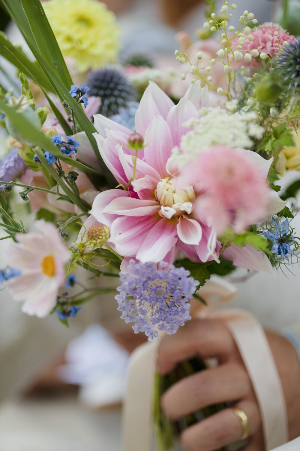 Close-up of a pastel wildflower bouquet with pink dahlias, purple scabiosa and blue blooms held in a hand wearing a gold ring.
