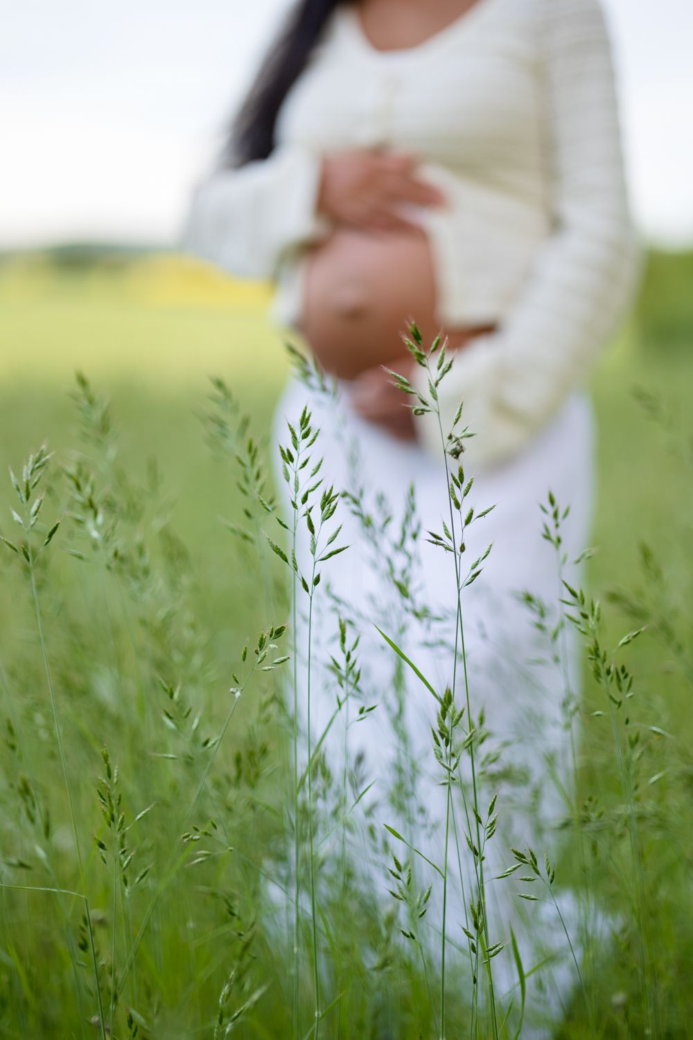 A pregnant woman in a cream cardigan and white skirt holds her belly in a green meadow with tall grass blurred in the foreground.