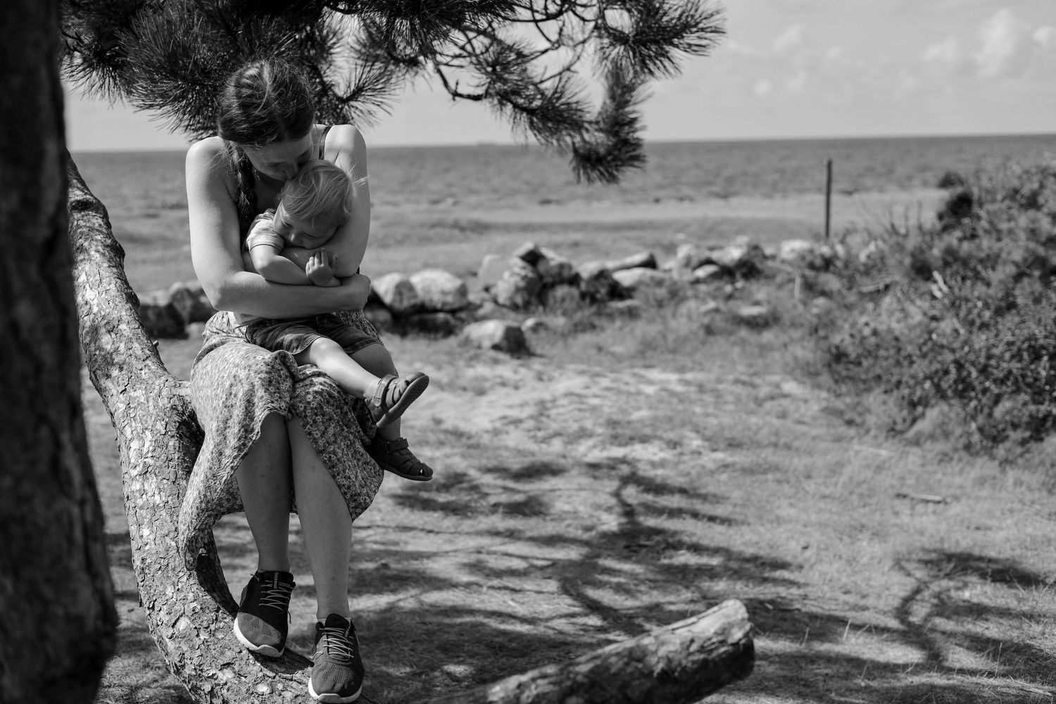 A black and white photo of a mother in a patterned dress sitting on a tree trunk holding her baby by the sea.