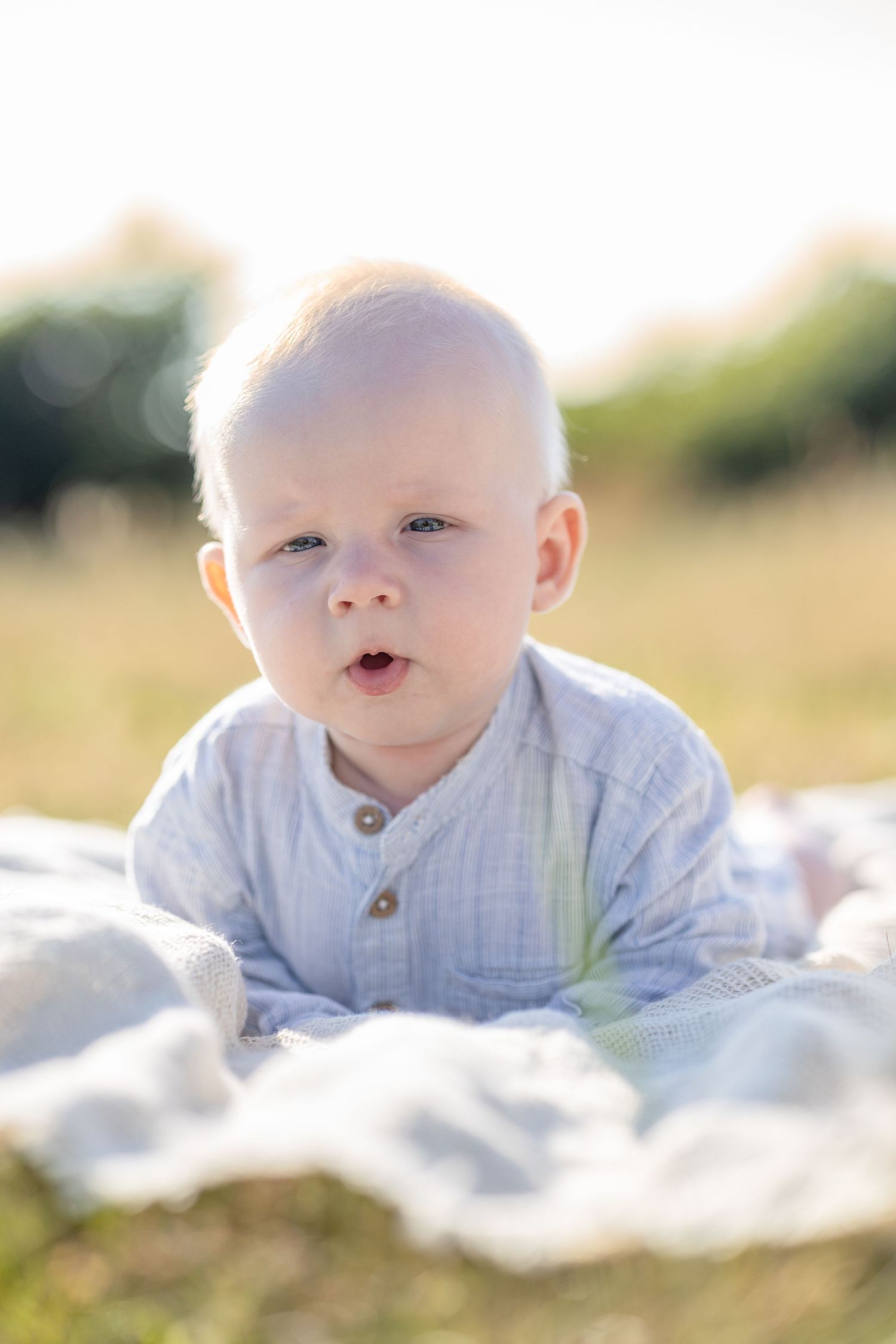 Smiling baby in a light blue button shirt lying on a soft blanket on a sunny grassy field.