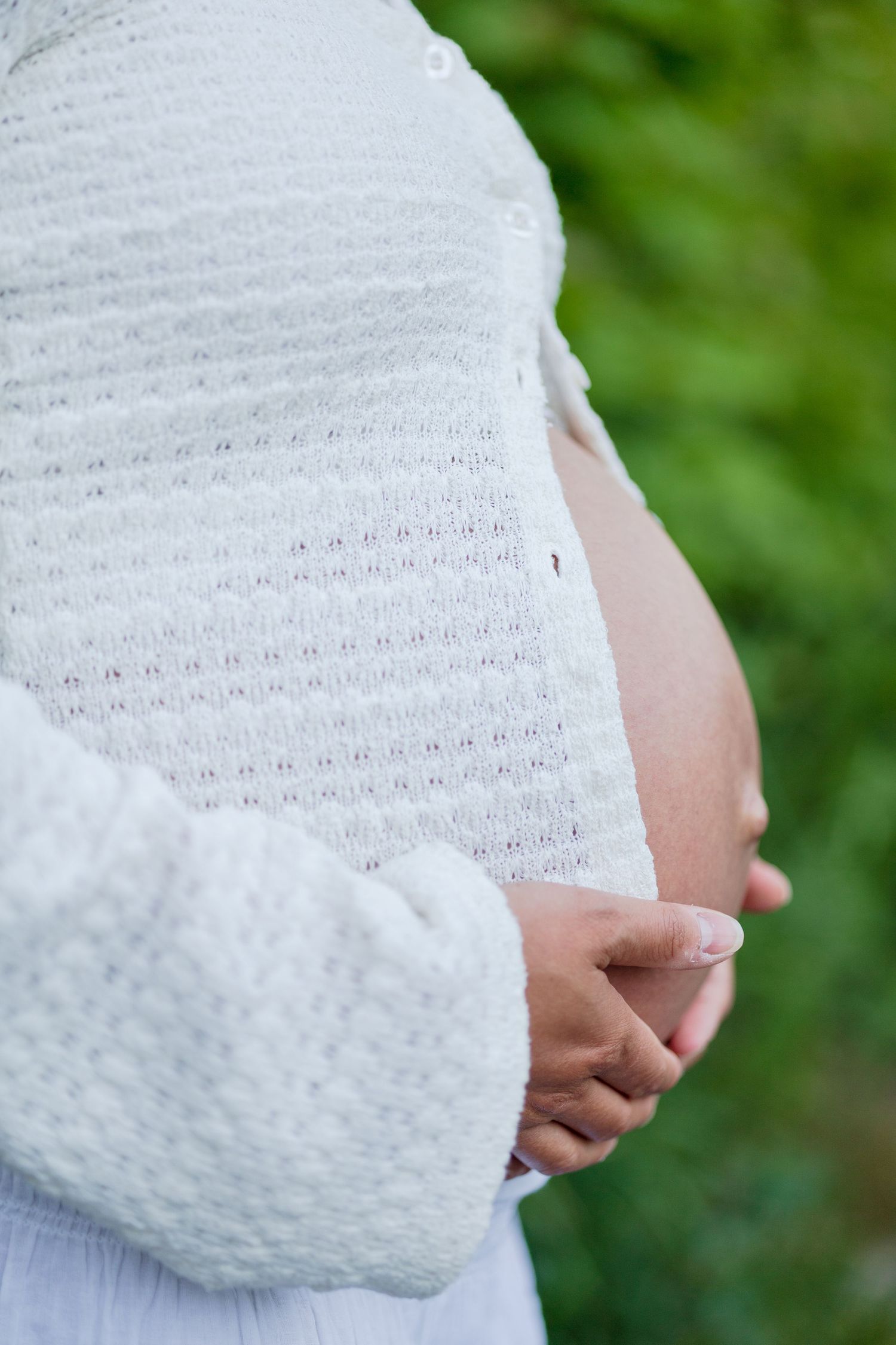 Close-up of a pregnant woman in a white knit cardigan gently holding her bare baby bump with both hands outdoors.