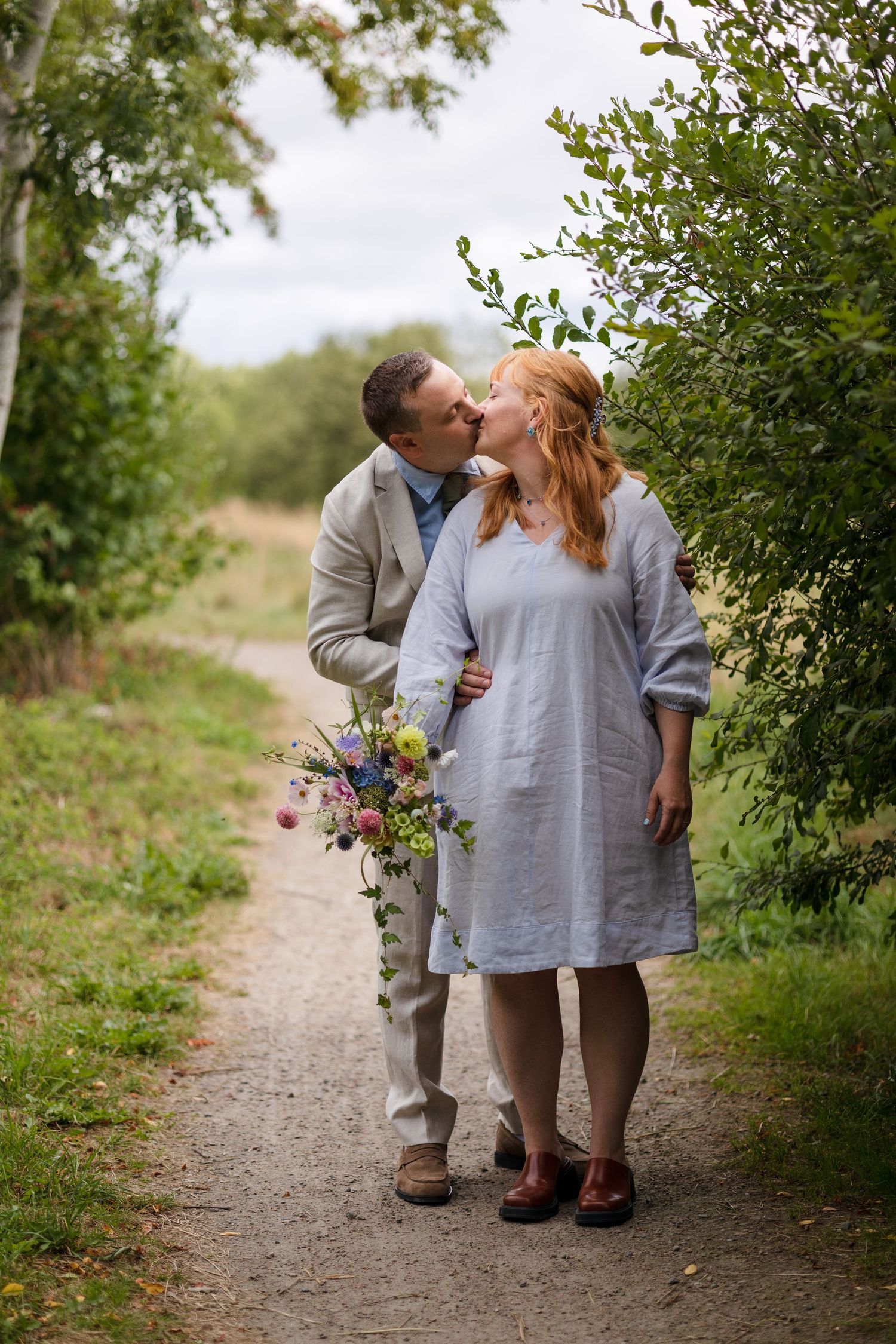 A couple shares a kiss on a gravel path lined with greenery, the woman holding a pink and purple wildflower bouquet.