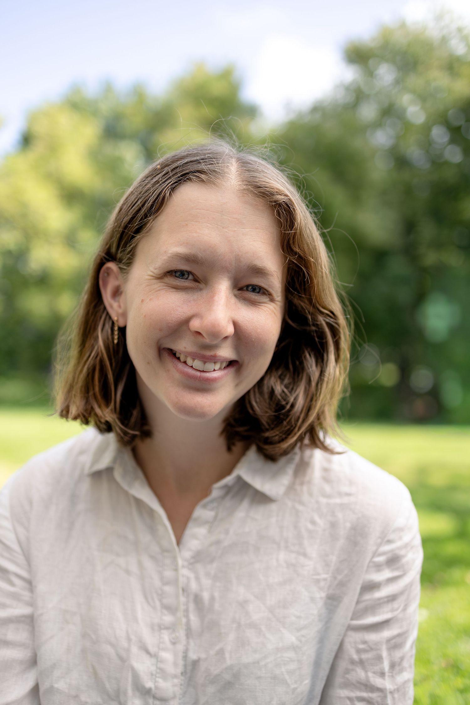 Smiling woman with shoulder-length brown hair wearing a white linen shirt standing in a sunny green park.
