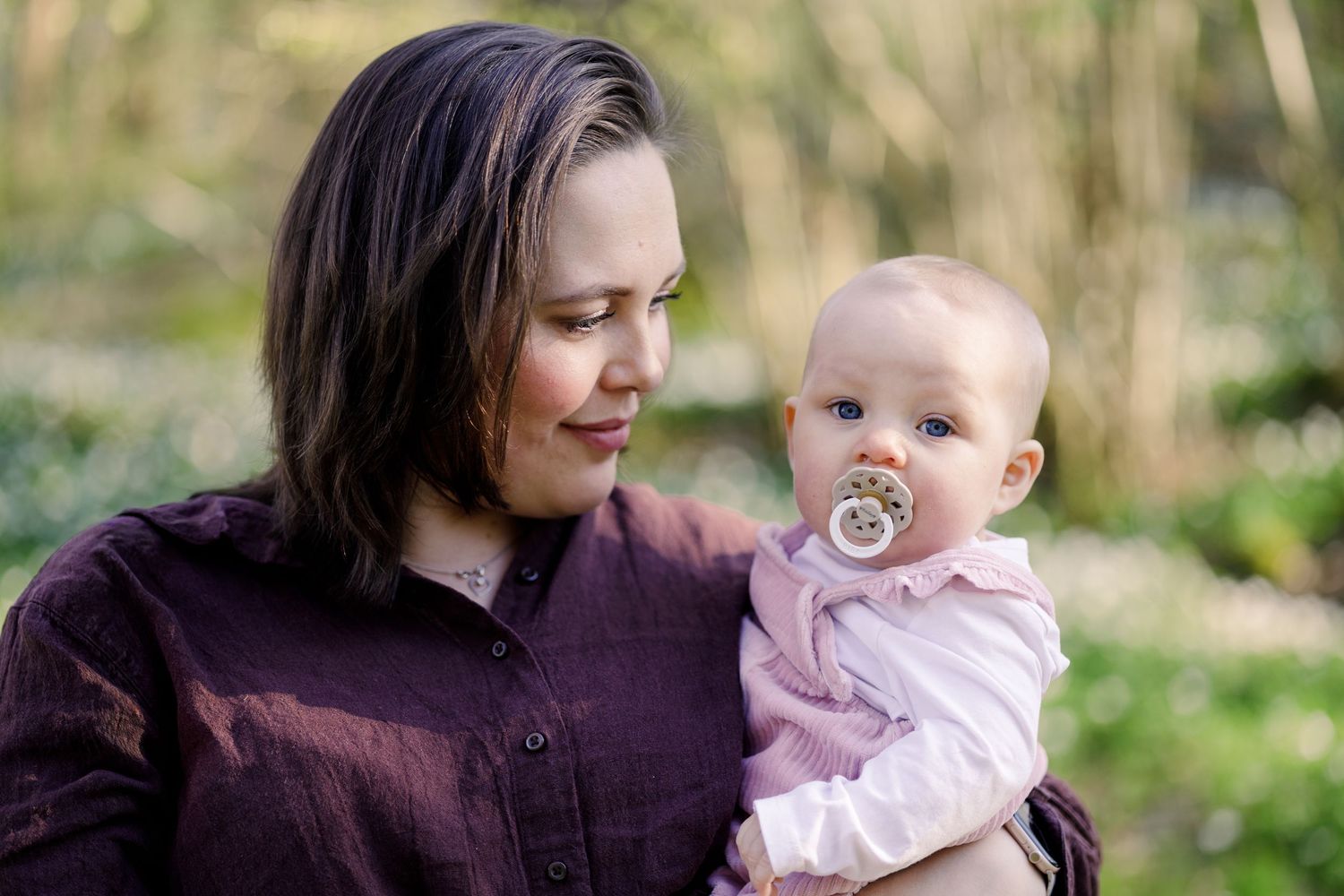 Smiling woman in a maroon shirt holds a baby with a pacifier in a soft-focus spring woodland setting.