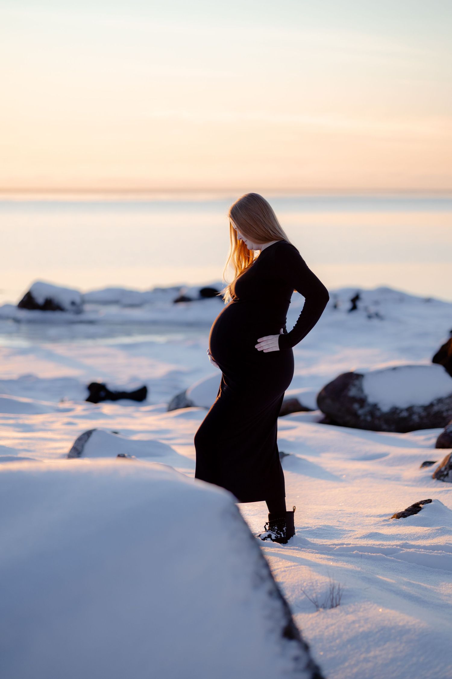 A pregnant woman in a black dress cradles her baby bump on a snowy shoreline during a soft pastel winter sunset.