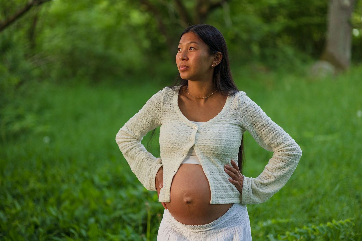 Pregnant woman in a cropped white cardigan rests her hands on her hips in a lush green park, gazing away.
