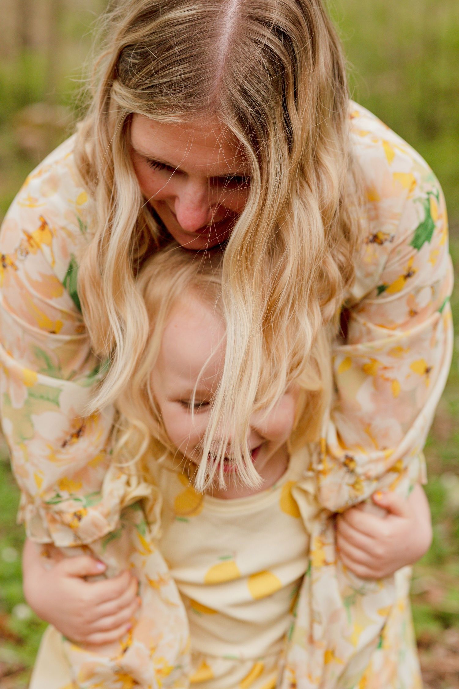 A woman with long blonde hair smiles down as a young child hugs her in a floral top outdoors.