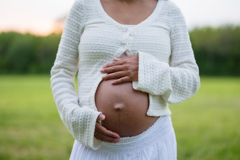 Close-up of a pregnant woman in a cream knit cardigan and white skirt cradling her bare belly in a green field.