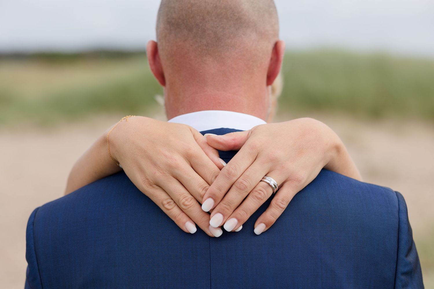 Bride's hands with a manicure and wedding ring wrap around the neck of a groom in a navy blue suit on a beach.