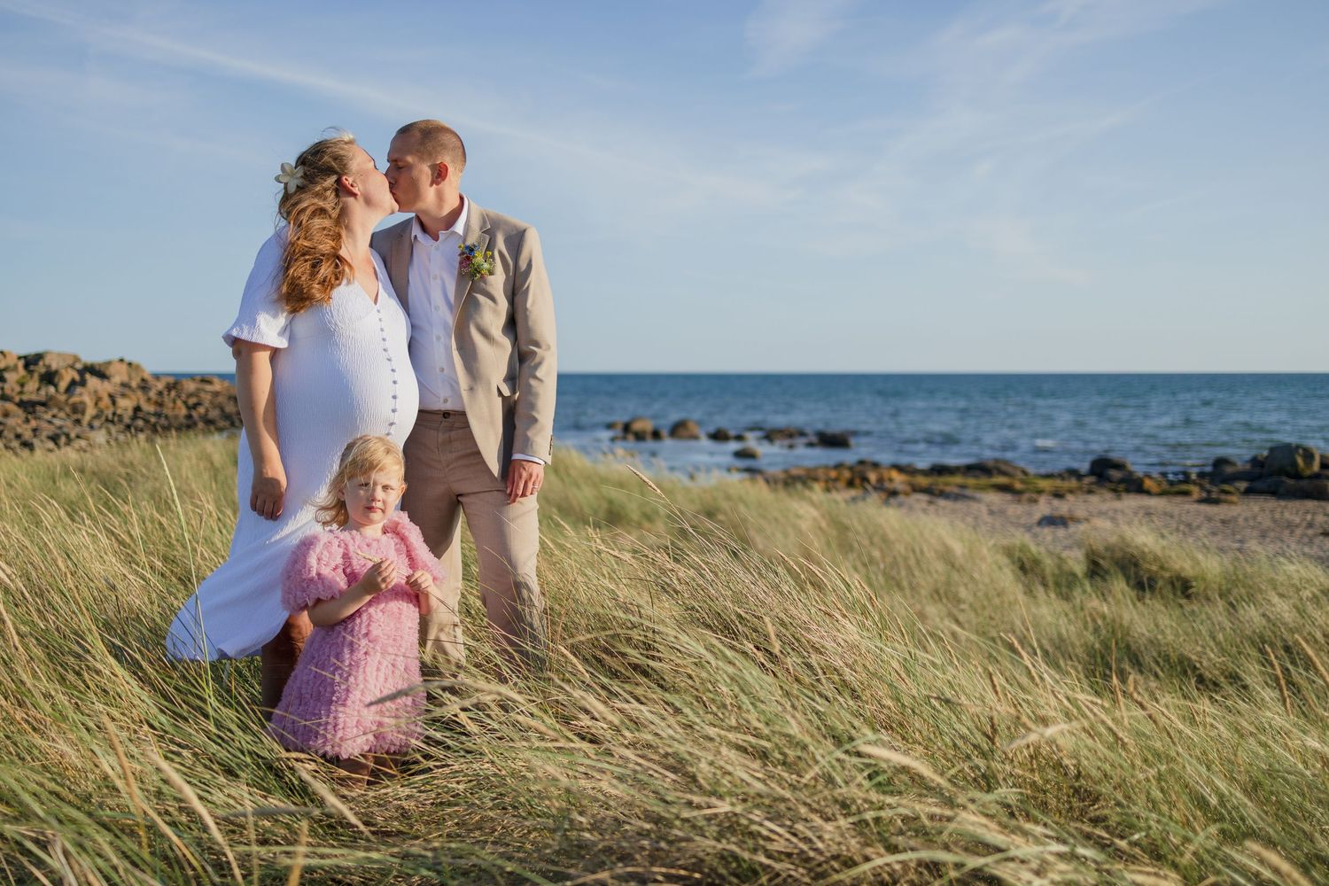 Bride and groom with a little girl in pink pose in tall beach grass with the sea behind them at sunset.