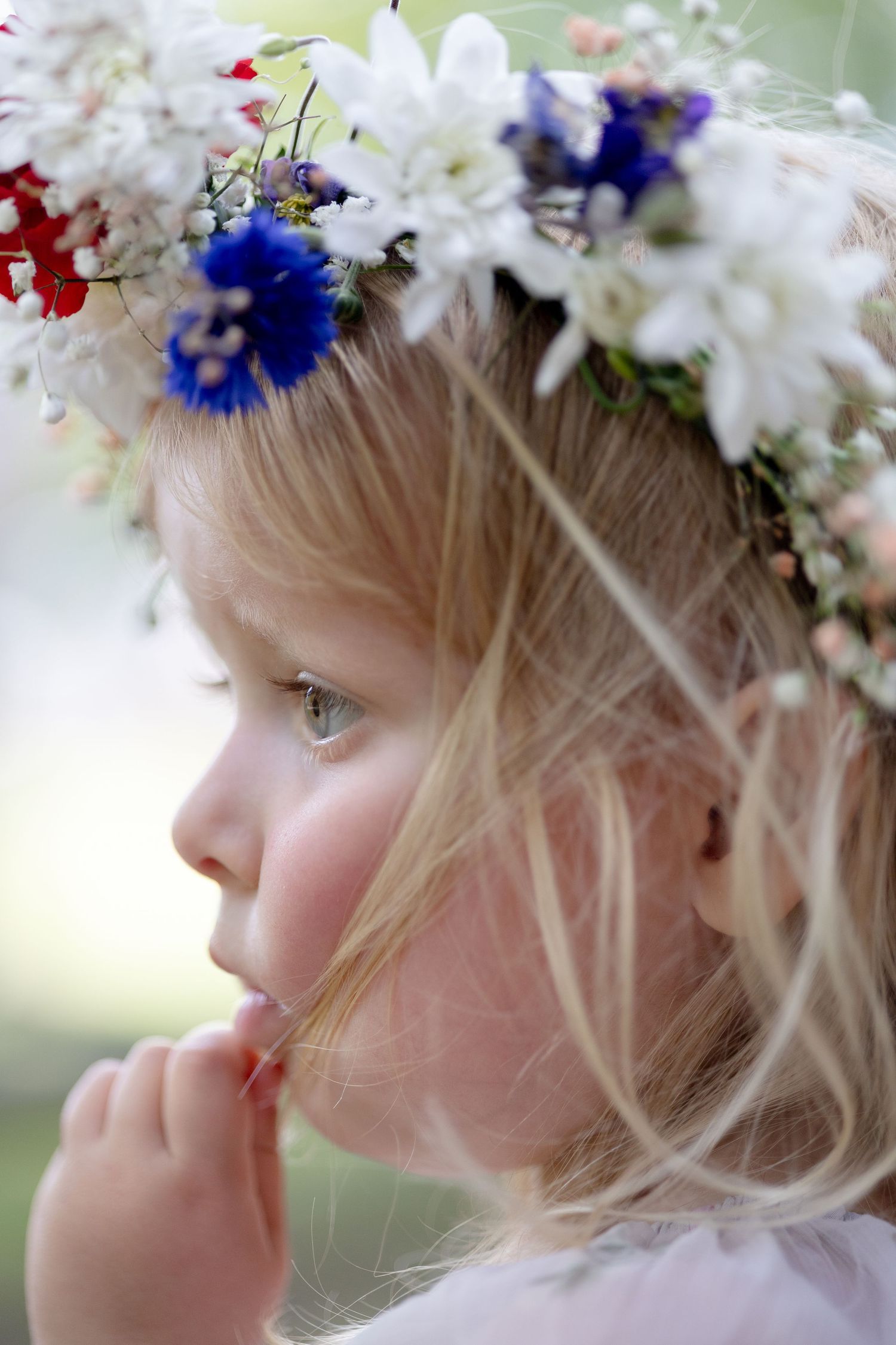 Profile of a young blonde girl wearing a flower crown of white, red and blue blooms while holding her hand to her mouth.
