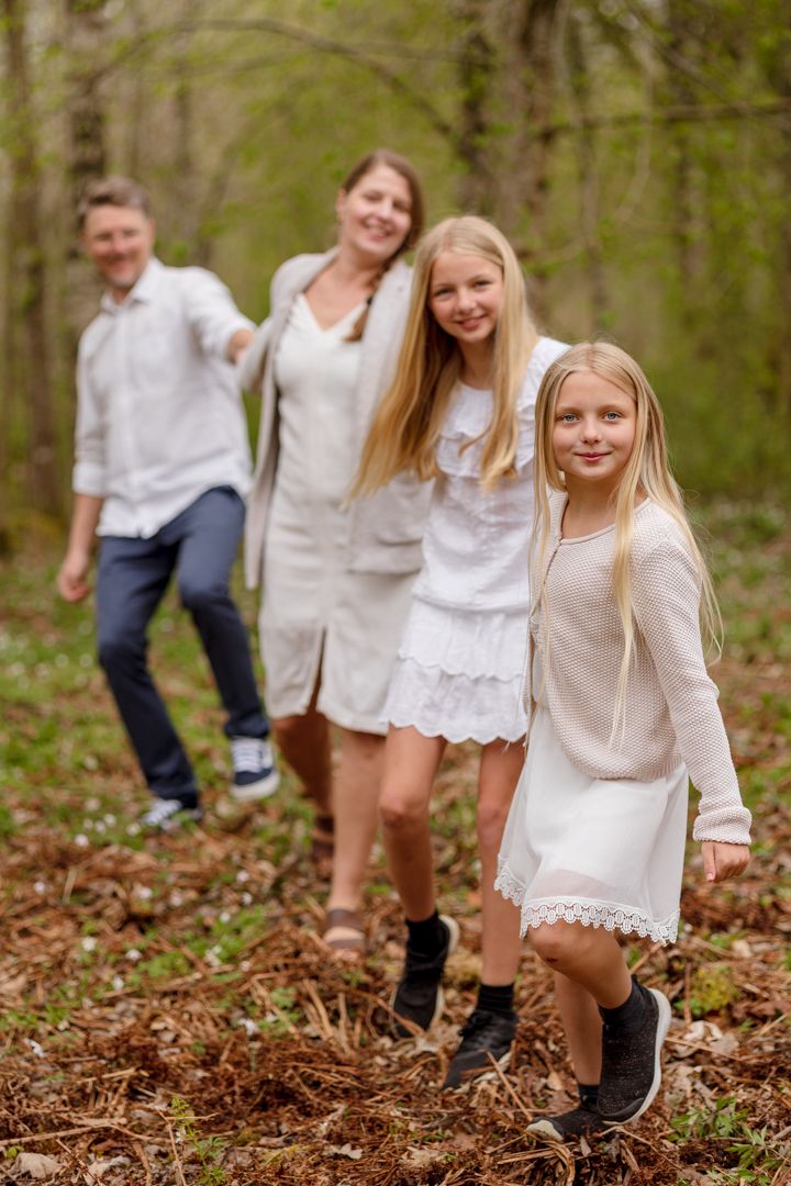 A smiling family of four in white and neutral outfits walks hand in hand through a green spring forest.